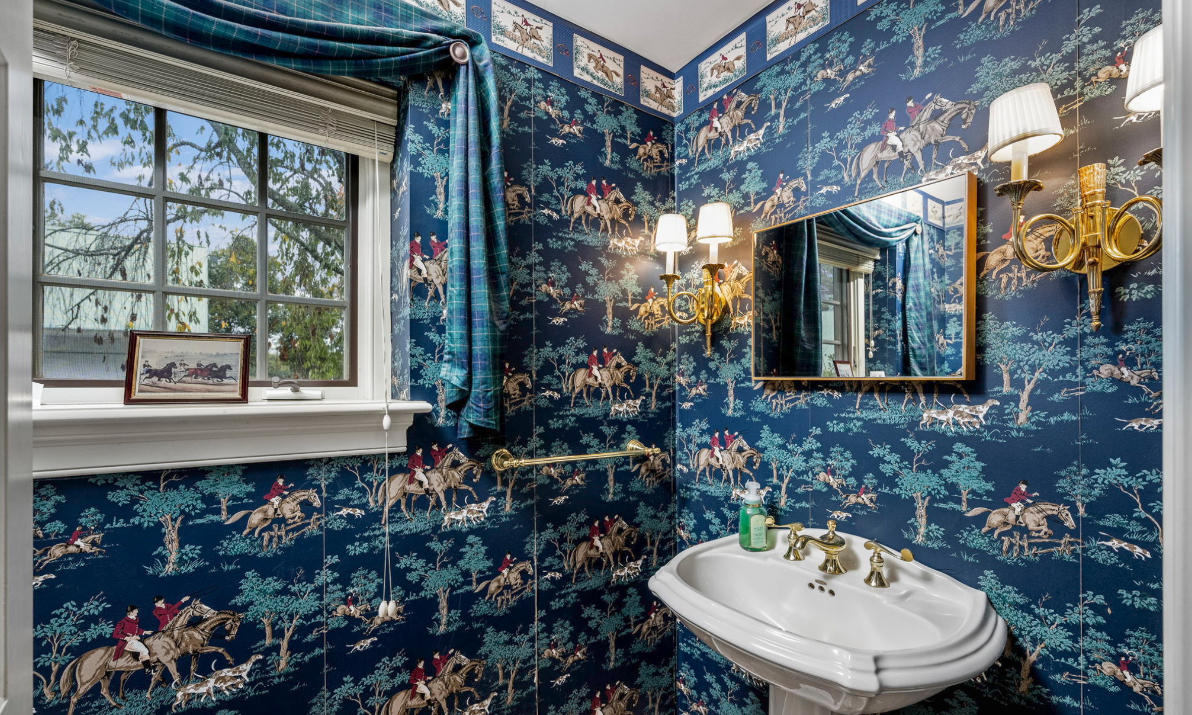 A bathroom with dark blue wallpaper featuring an intricate pattern of people riding horses through a forest, with framed pictures on the wall and a mirror above the sink.