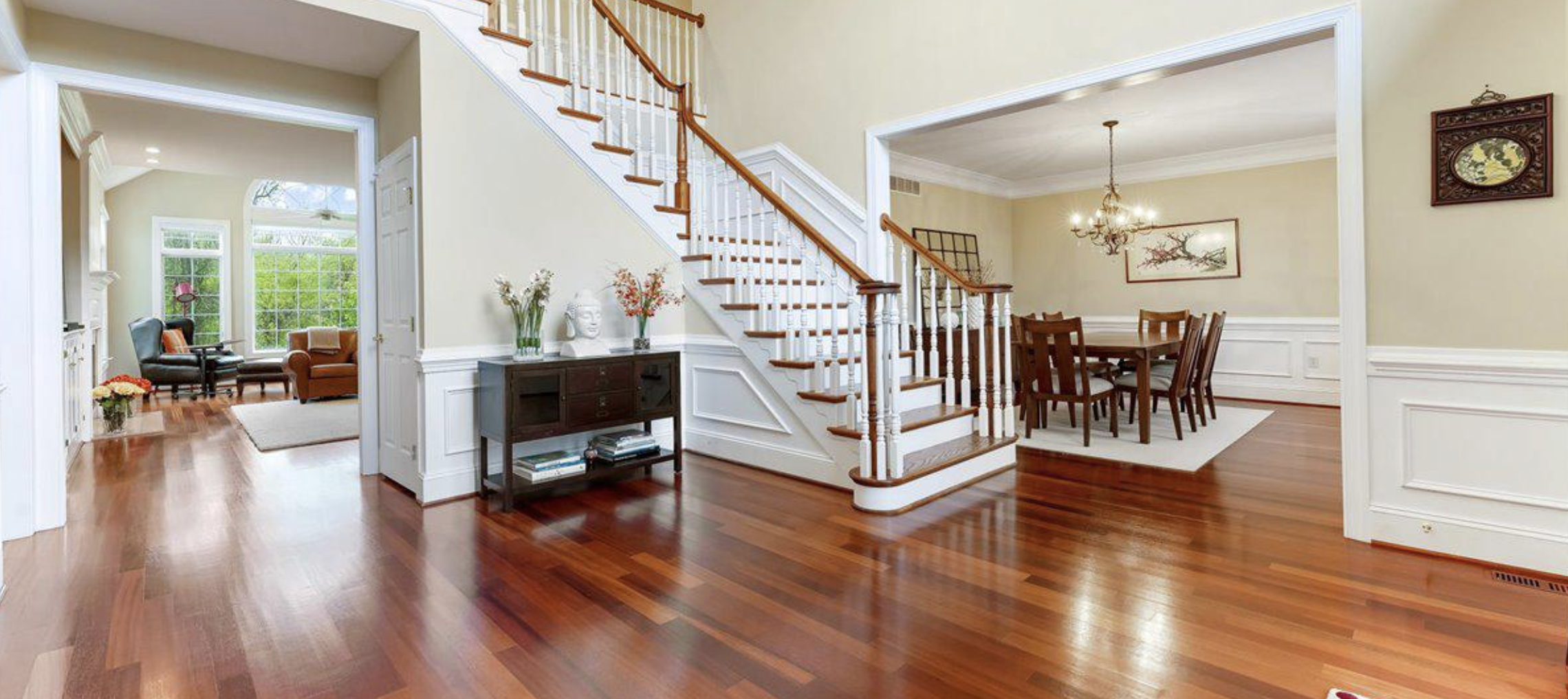 Interior view of a house with hardwood floors, featuring a staircase with white spindles and wooden handrails, a black console table with decorative items, and a dining area with a wooden table and chairs. There is a living room in the background with large windows and seating.