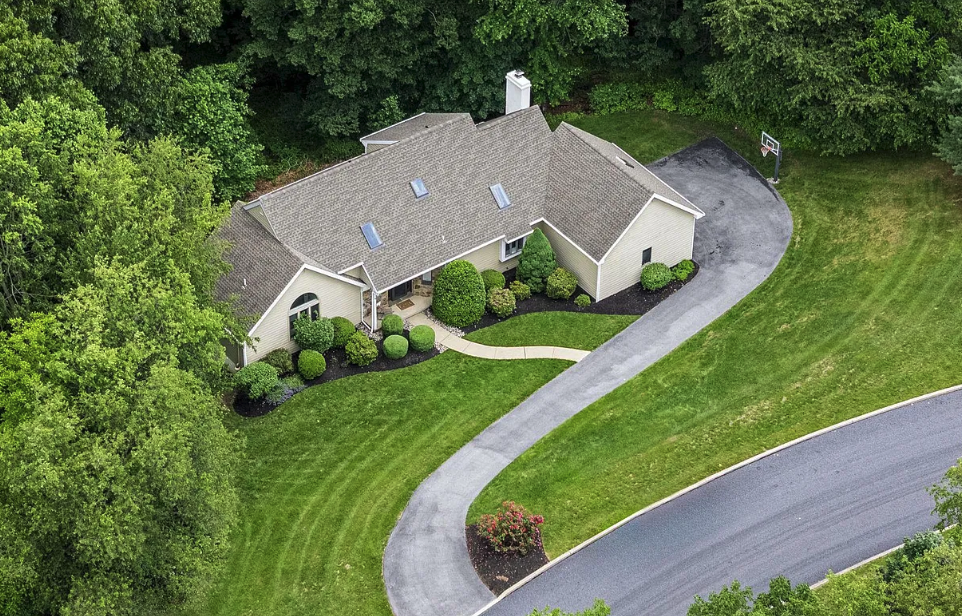 Aerial view of a single-story house with a gray roof, surrounded by a well-maintained lawn, trees, bushes, a curved driveway, and a basketball hoop in the backyard.