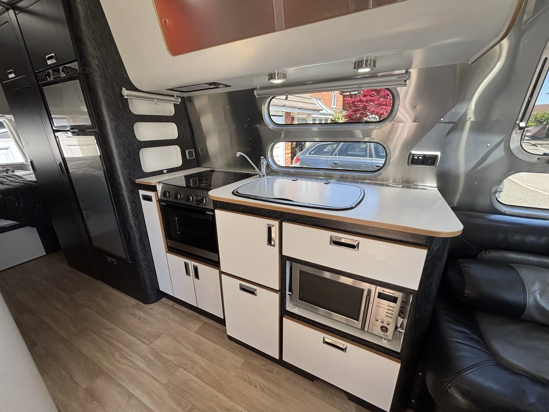 Interior of a compact camper van with kitchen area and black leather seating, window view of parked cars and a red-leafed tree outside.