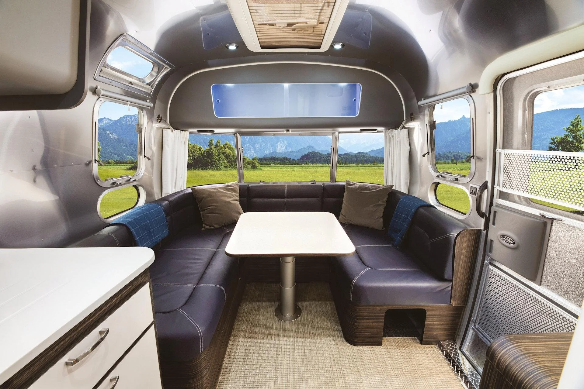 Interior of a retro-style camper van with large windows showing a mountain landscape, a curved black bench with cushions, a white table, and modern amenities.