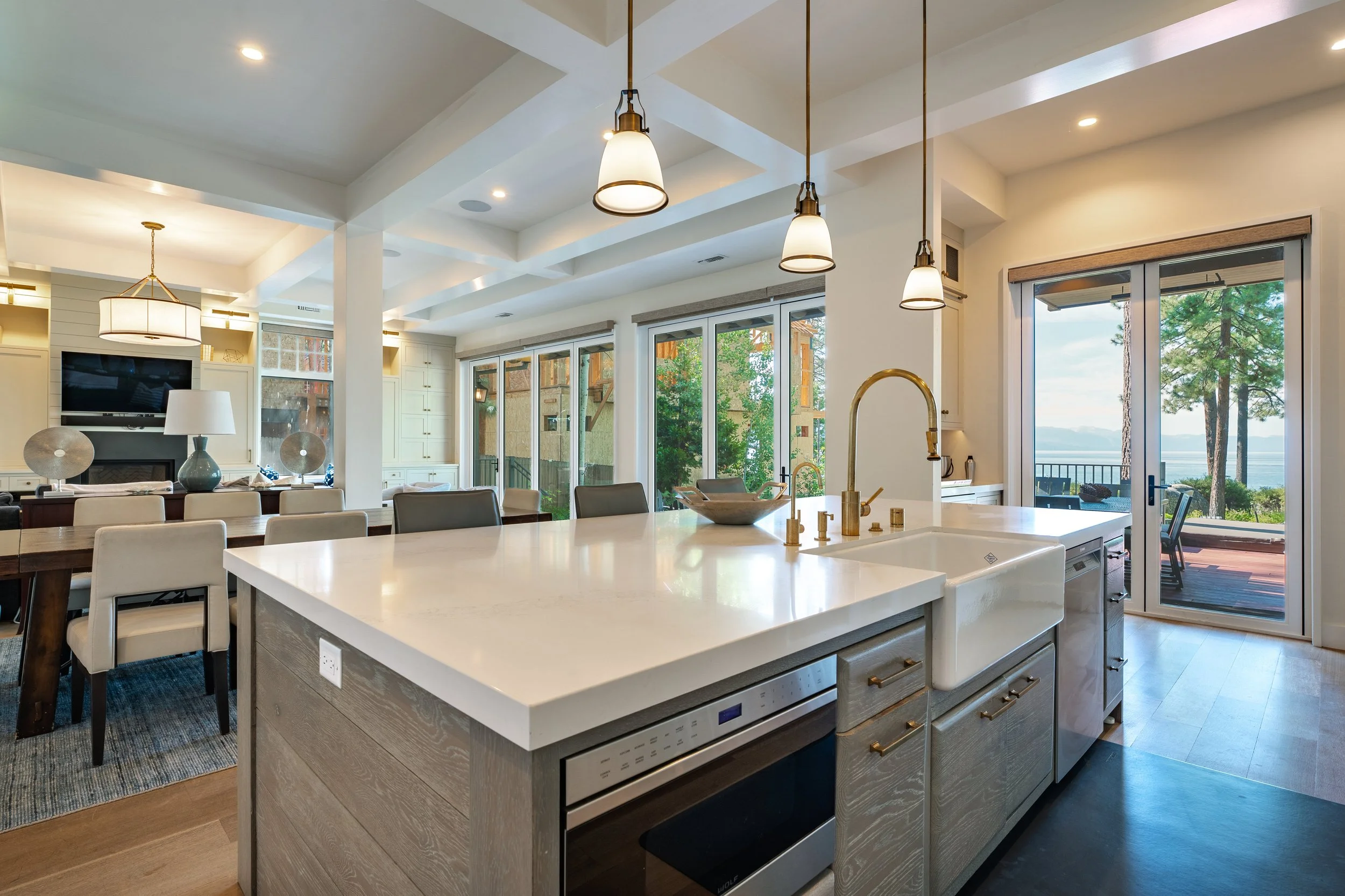 Open-concept kitchen with white island, gold pendant lights, and large windows showing trees and a lake view outside.