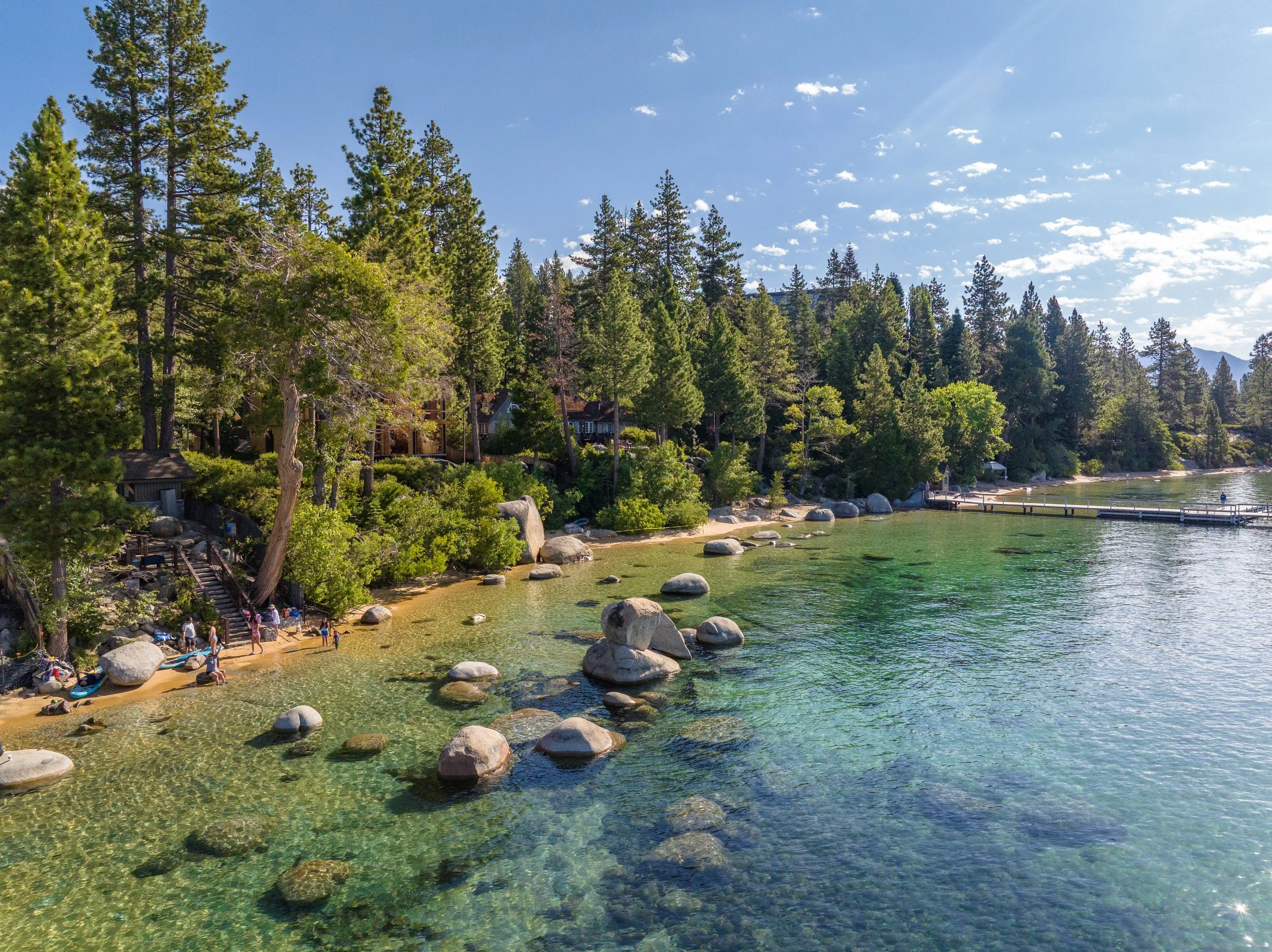 A lakeside scene with a sandy beach, large rocks in the shallow water, and a dense forest of tall pine trees in the background under a bright blue sky with scattered clouds.