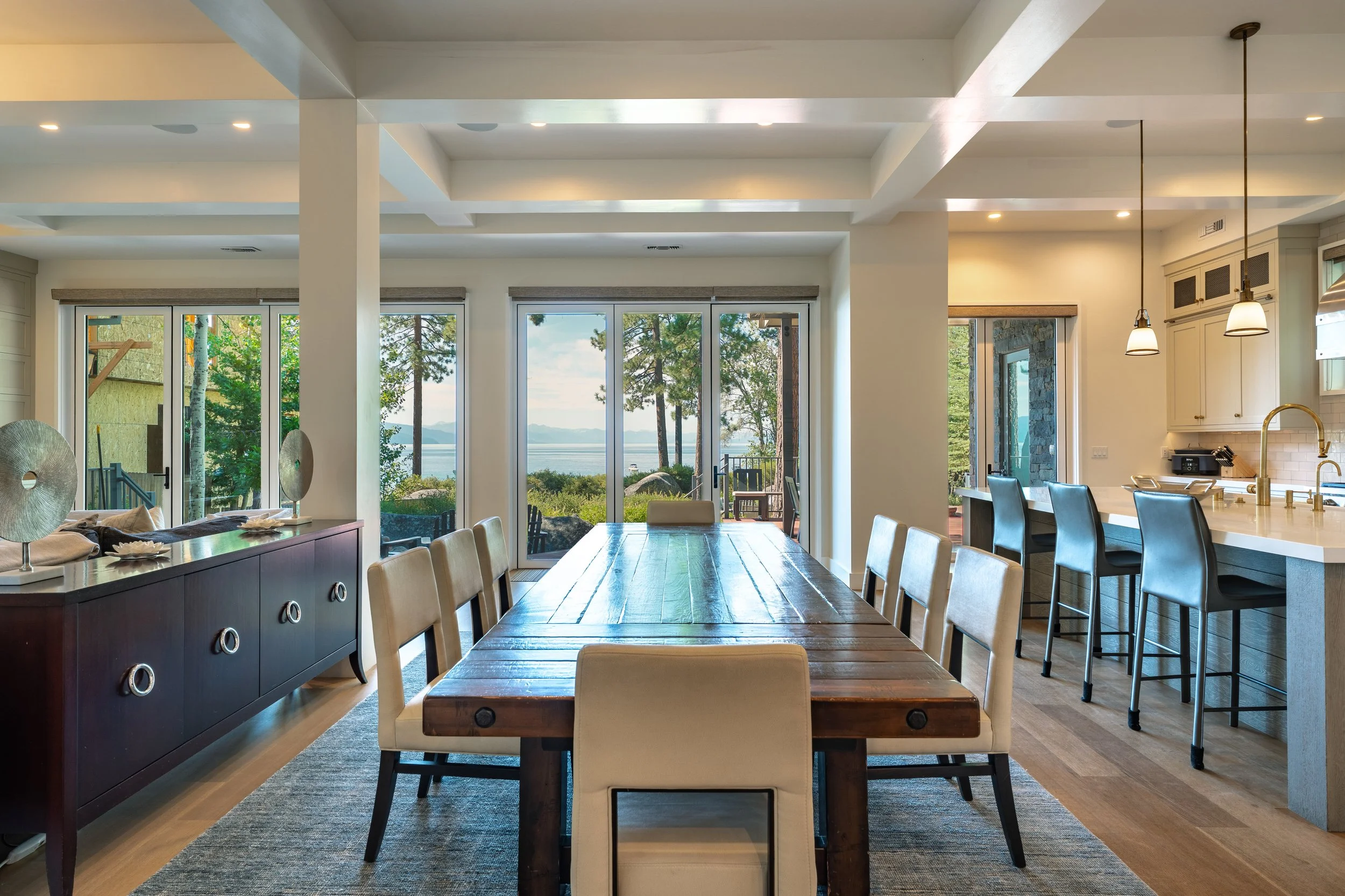 Open-concept living and dining area with large glass sliding doors overlooking a lake and trees, with kitchen on the right.