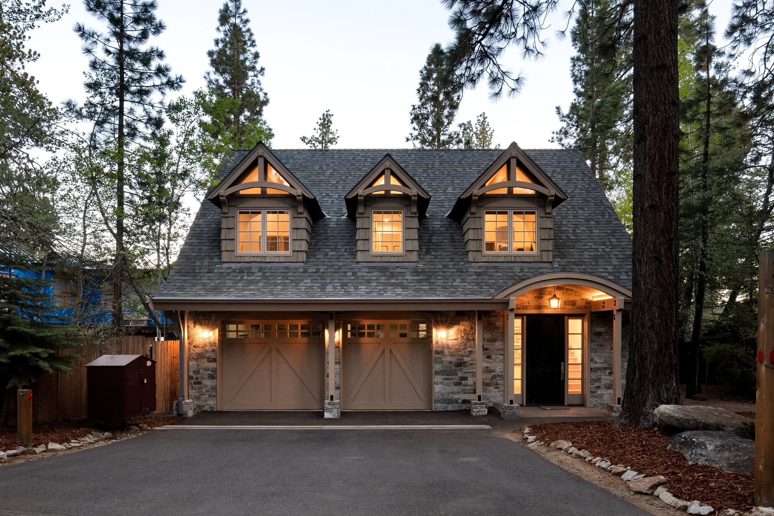 A two-story house in a wooded area with a peaked roof, three dormer windows, and an illuminated garage and front door.