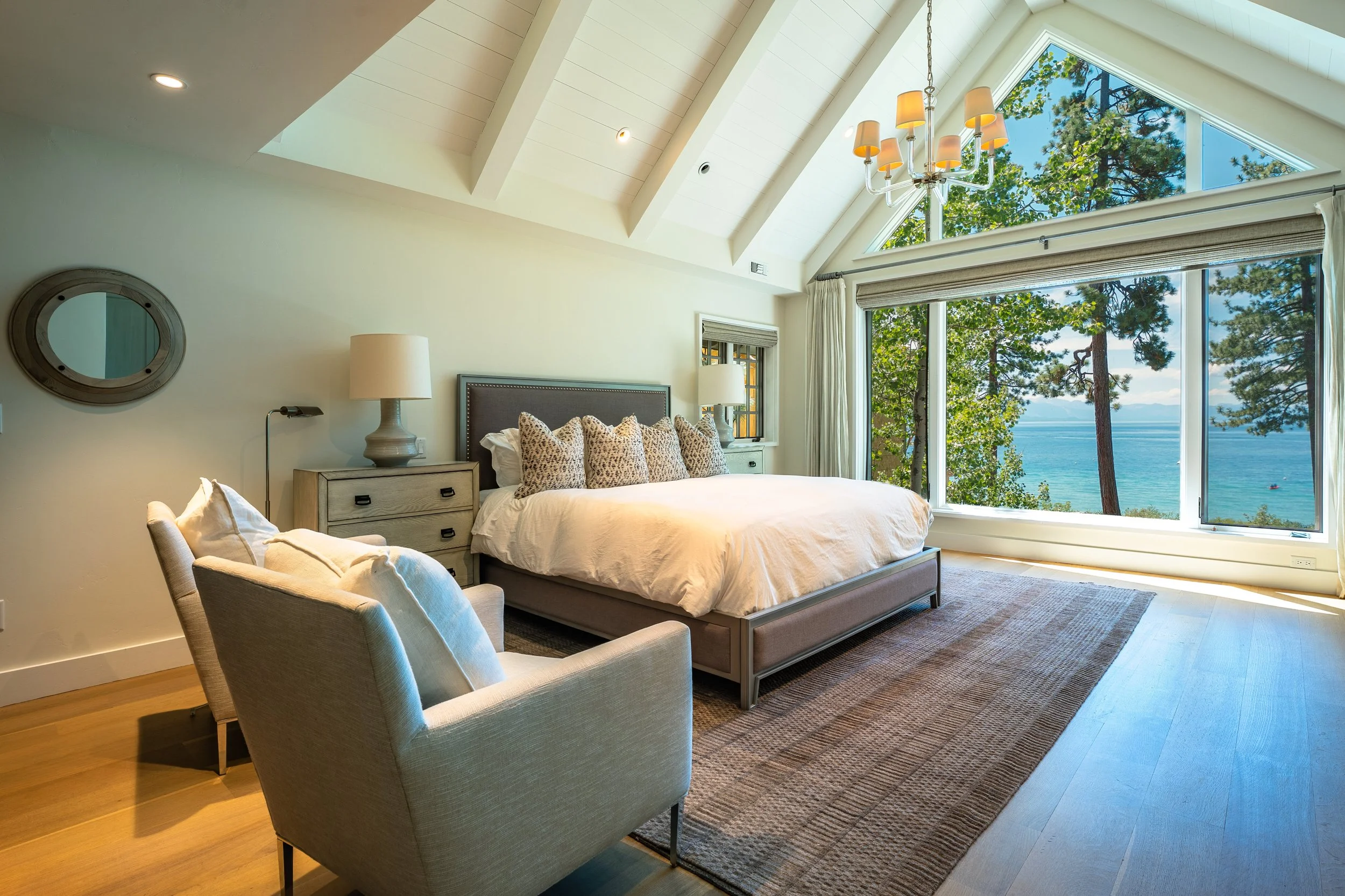 A modern bedroom with large windows showcasing a scenic view of trees and water. The room includes a bed with leopard-print pillows, nightstands with lamps, a cozy armchair, and a contemporary chandelier hanging from the angled ceiling.