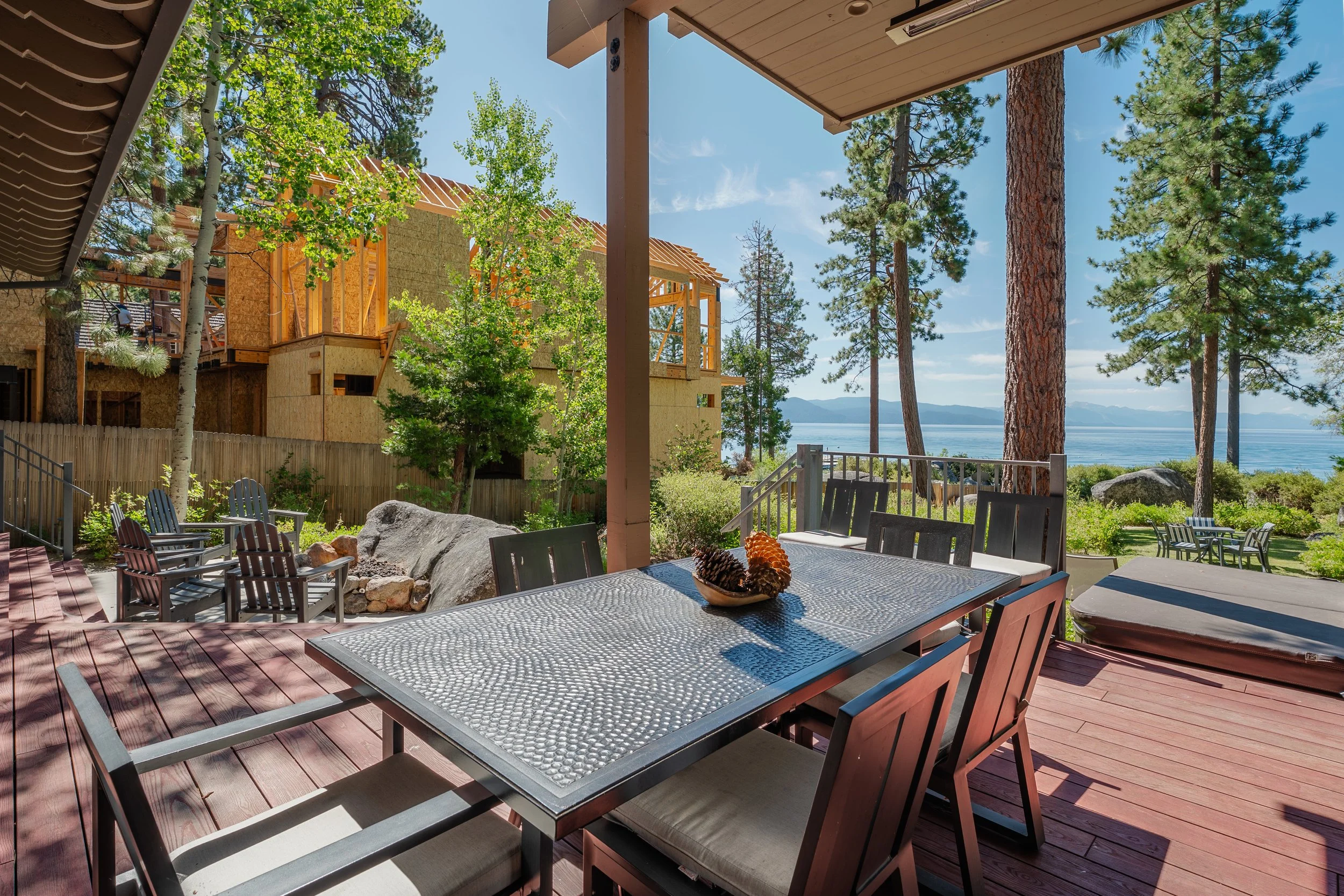 Outdoor patio with a large metal dining table, chairs, and a view of trees, water, and distant mountains on a bright, sunny day.