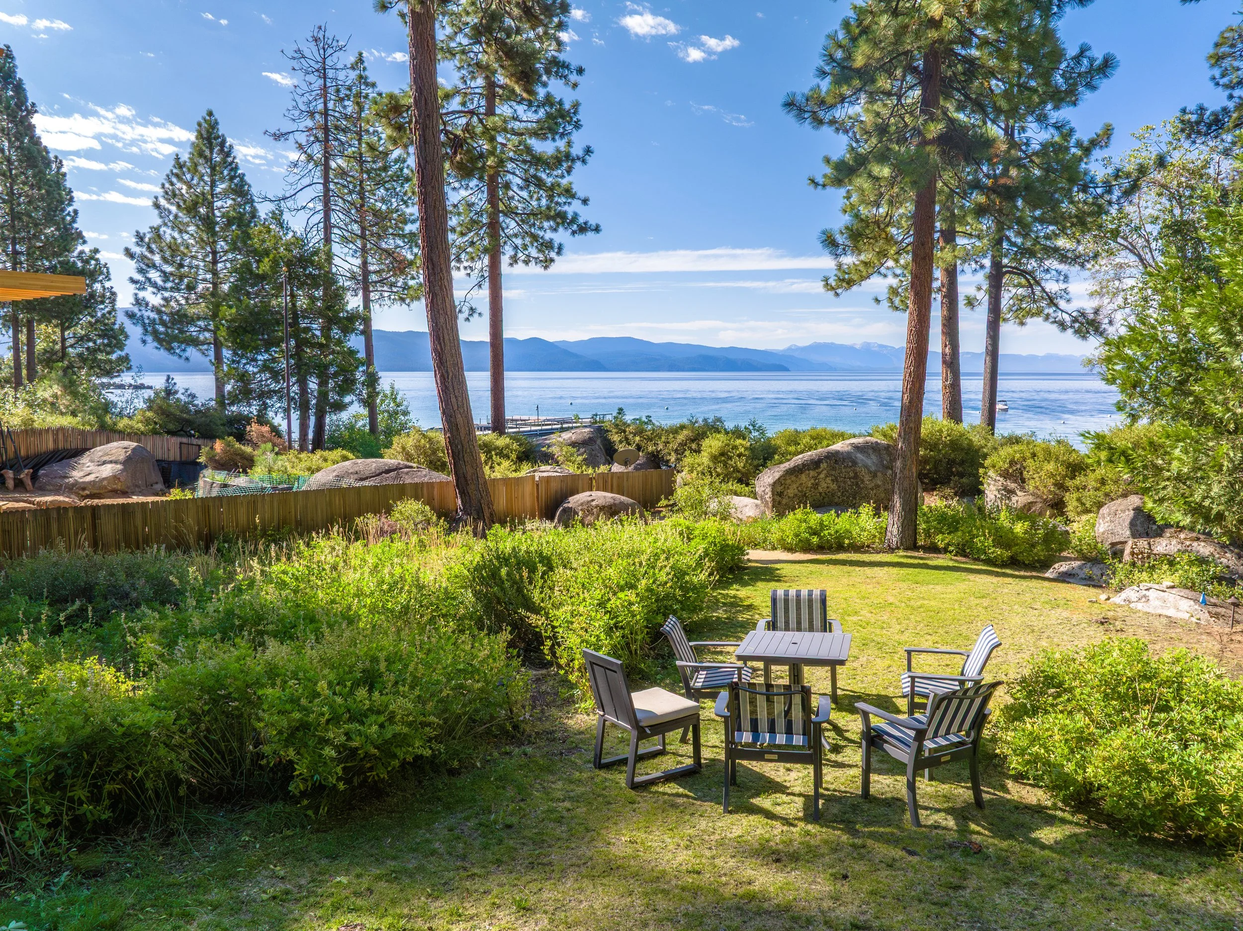 A backyard garden with outdoor chairs, lush green plants, tall pine trees, and a view of a large body of water with mountains in the background under a partly cloudy blue sky.