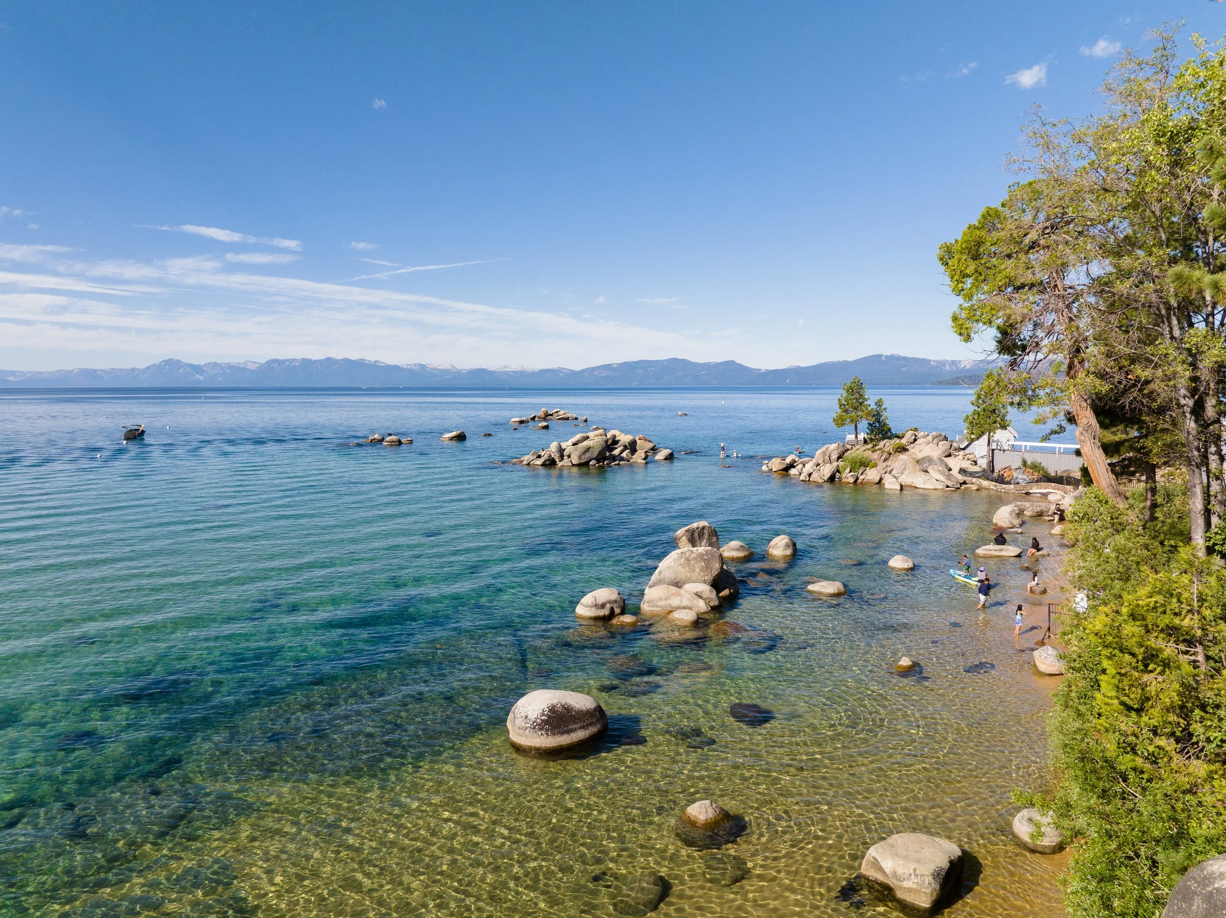 Clear lake with colorful water, rocks along the shoreline, trees on the right, and mountains in the background under a blue sky with a few clouds.