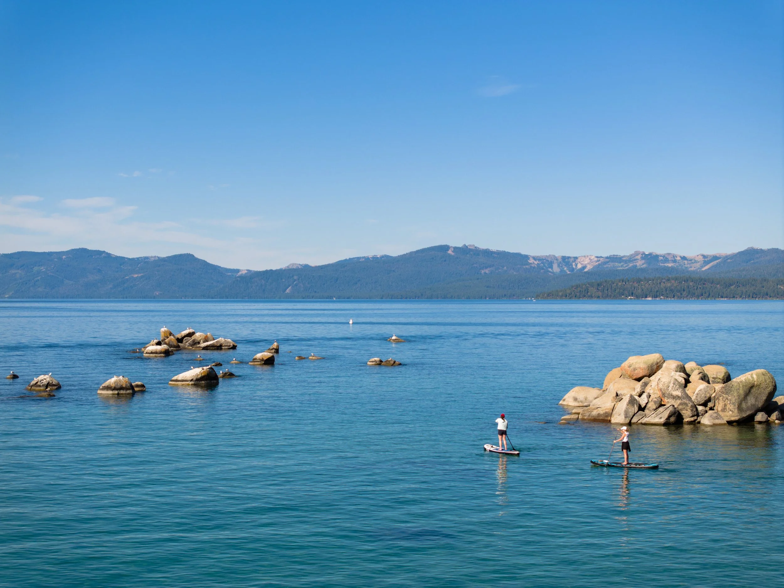 People paddleboarding and relaxing by the water with mountains in the background.