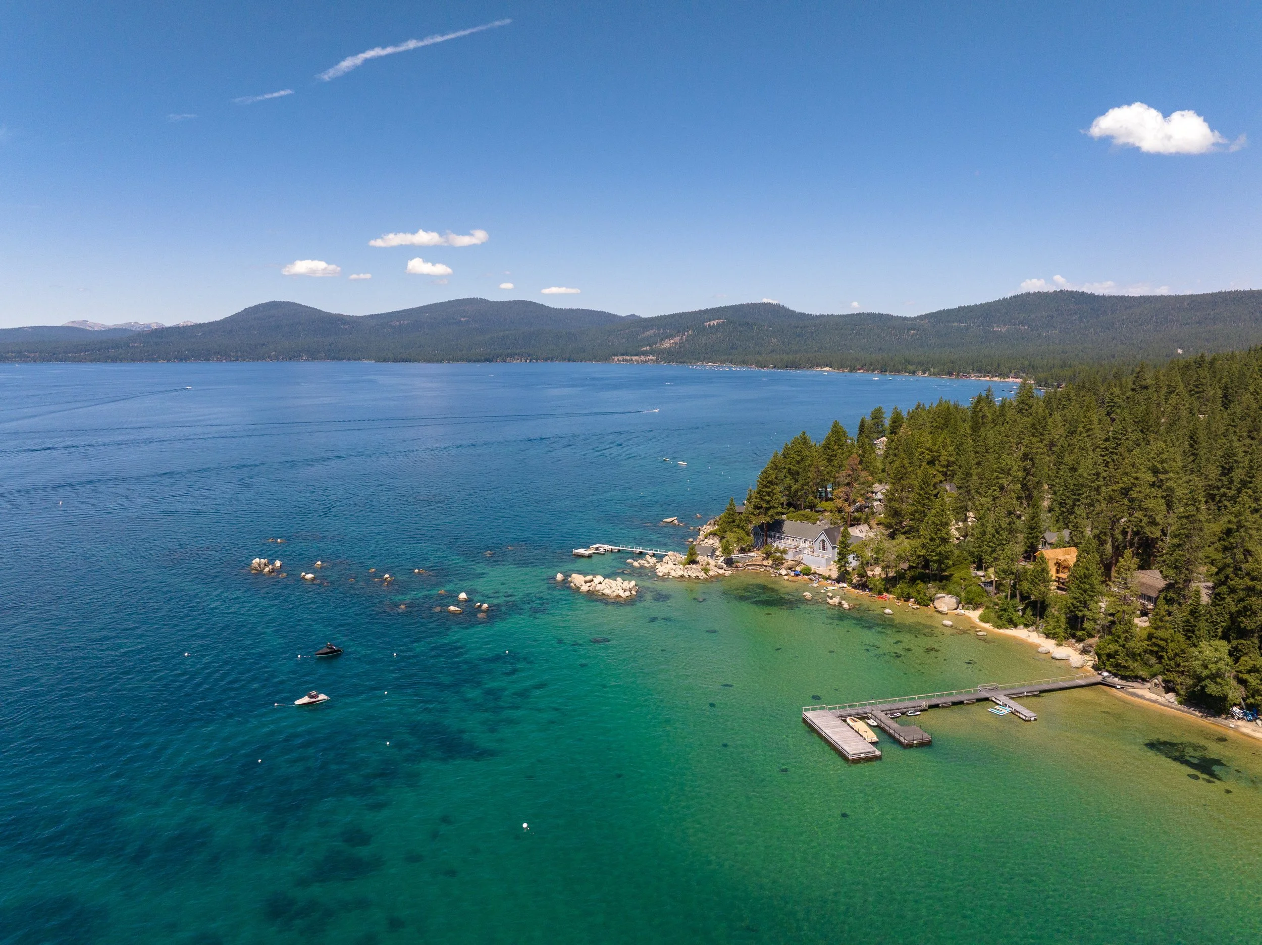 Aerial view of a large lake with a shoreline wooded with pine trees, houses, docks, and boats on the water, with mountains in the background under a clear blue sky with a few clouds.
