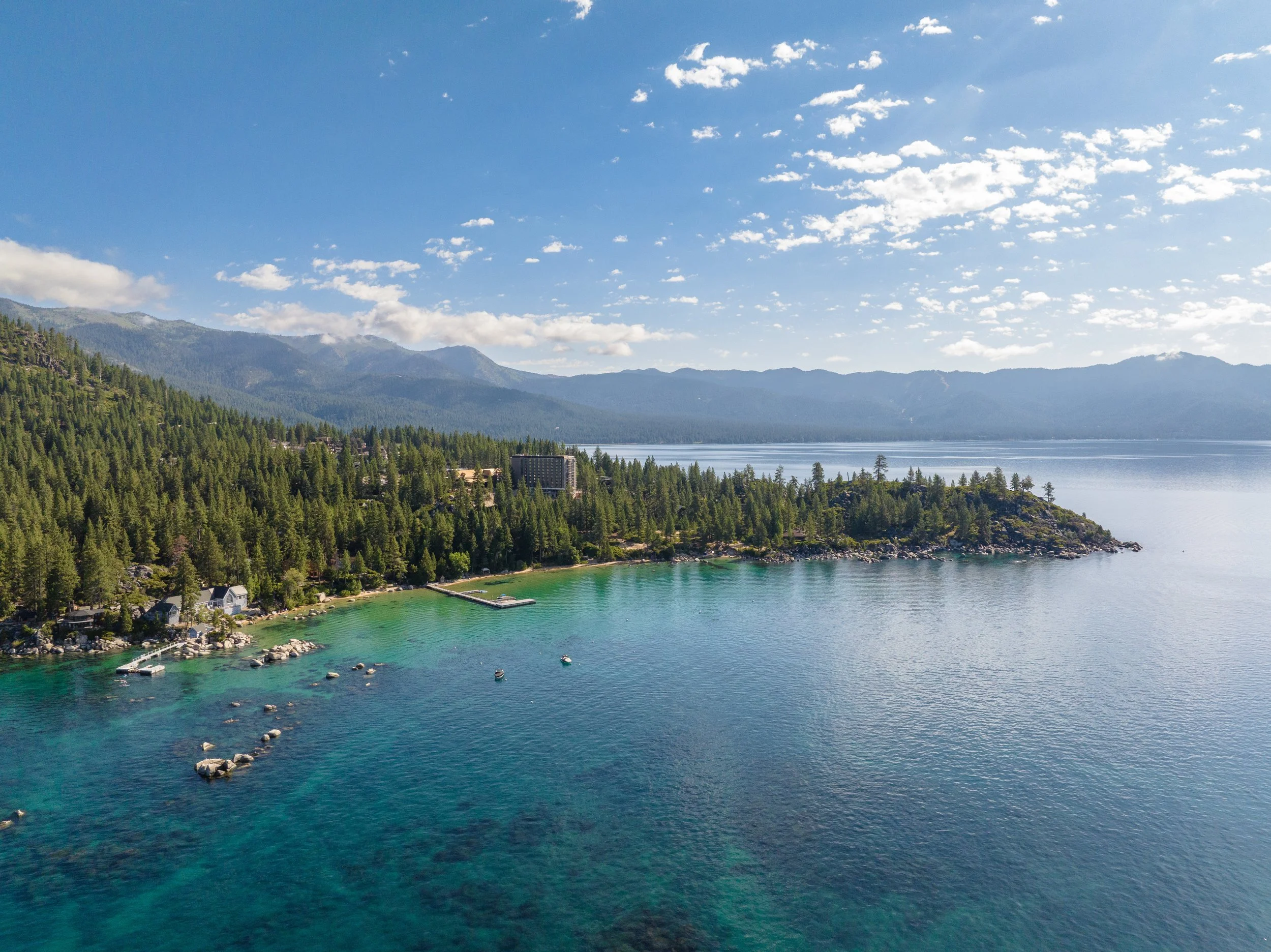 A scenic view of a lake with turquoise water, surrounded by dense pine forests, with mountains in the background under a blue sky filled with scattered clouds.