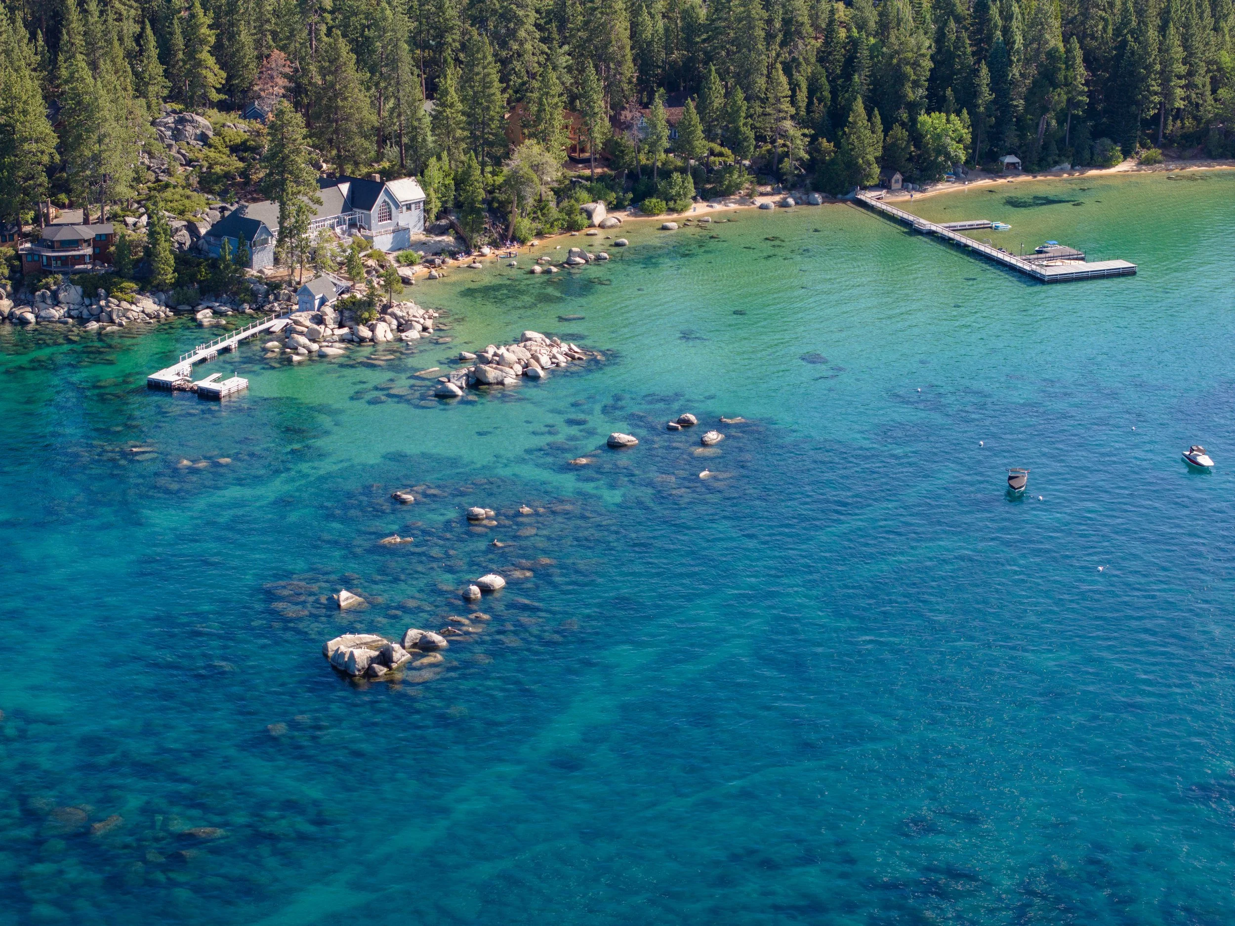 An aerial view of a lakeside area with houses, a sandy beach, a dock extending into the water, and boats floating on the water. The shoreline is lined with trees and large rocks.