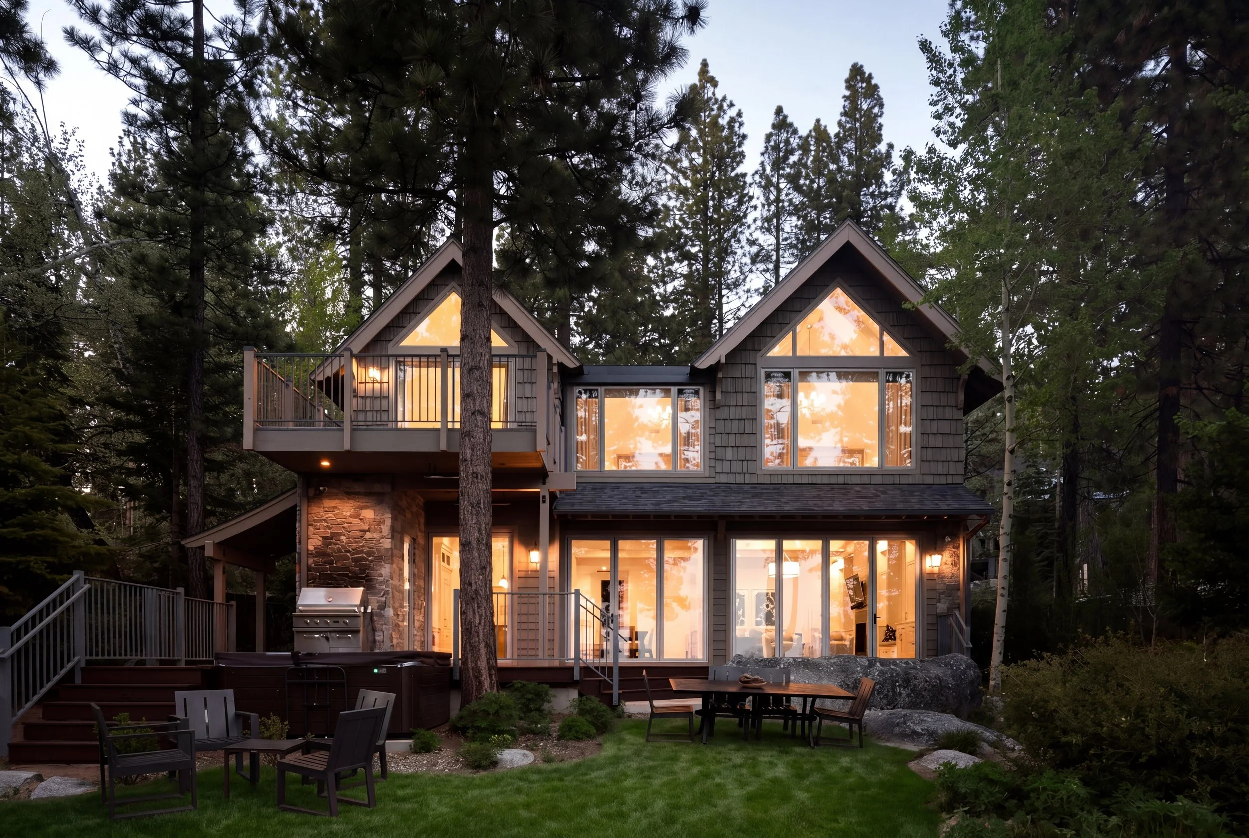 Modern house with large glass windows illuminated from inside, surrounded by tall trees and a lush green yard with outdoor furniture and a grill.