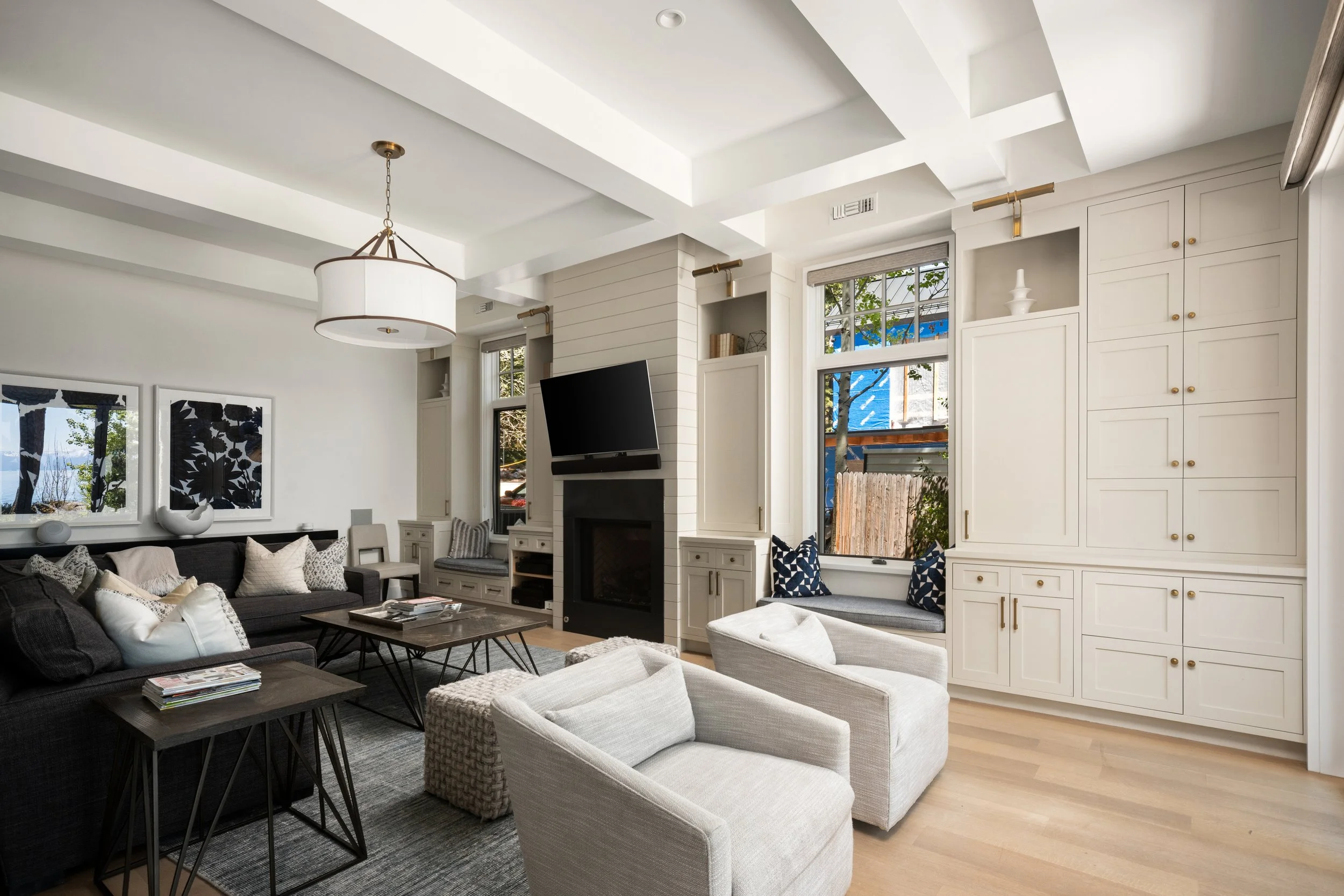 Living room with white coffered ceiling, large windows, white built-in cabinets, black and white sofas, coffee tables, and decorative pillows.