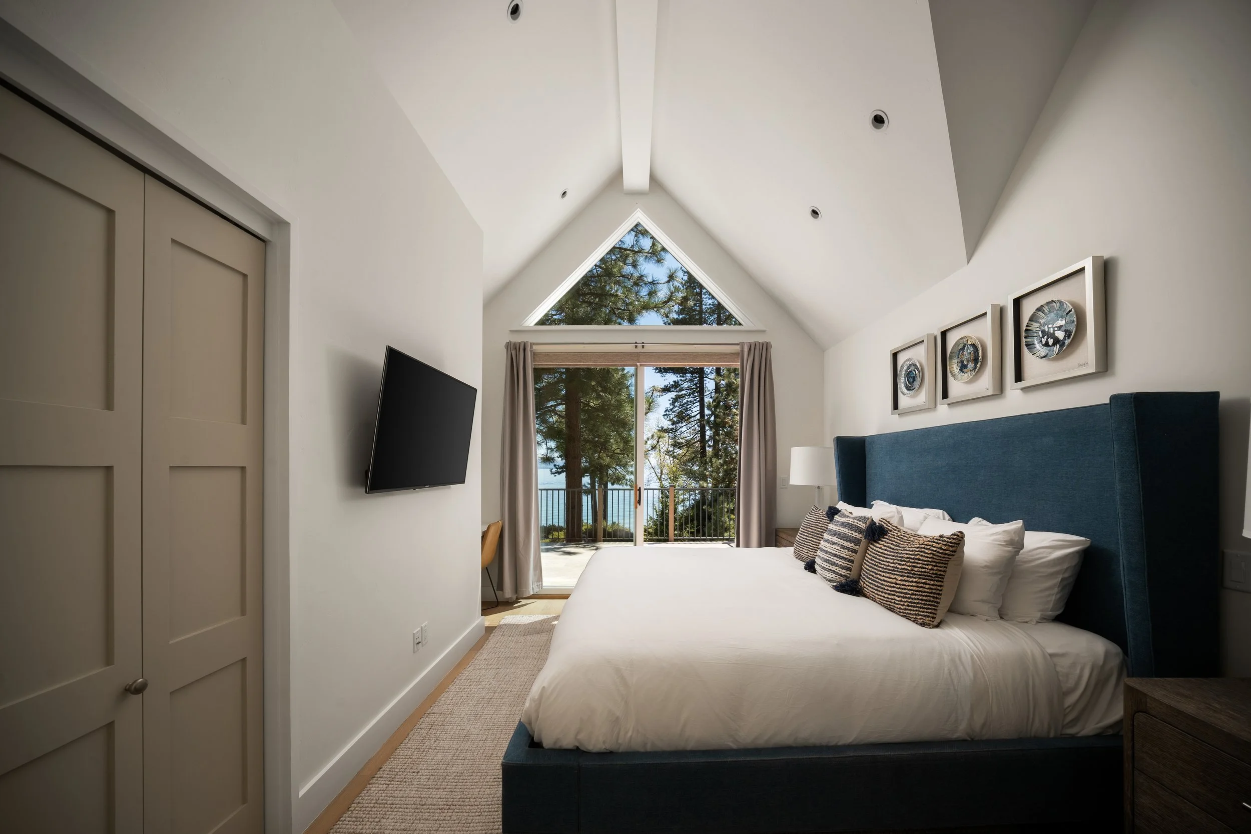 A bedroom with a large window showing trees outside. The room has a blue upholstered bed with multiple decorative pillows. There are framed pictures on the wall and a flat-screen TV mounted beside the window.
