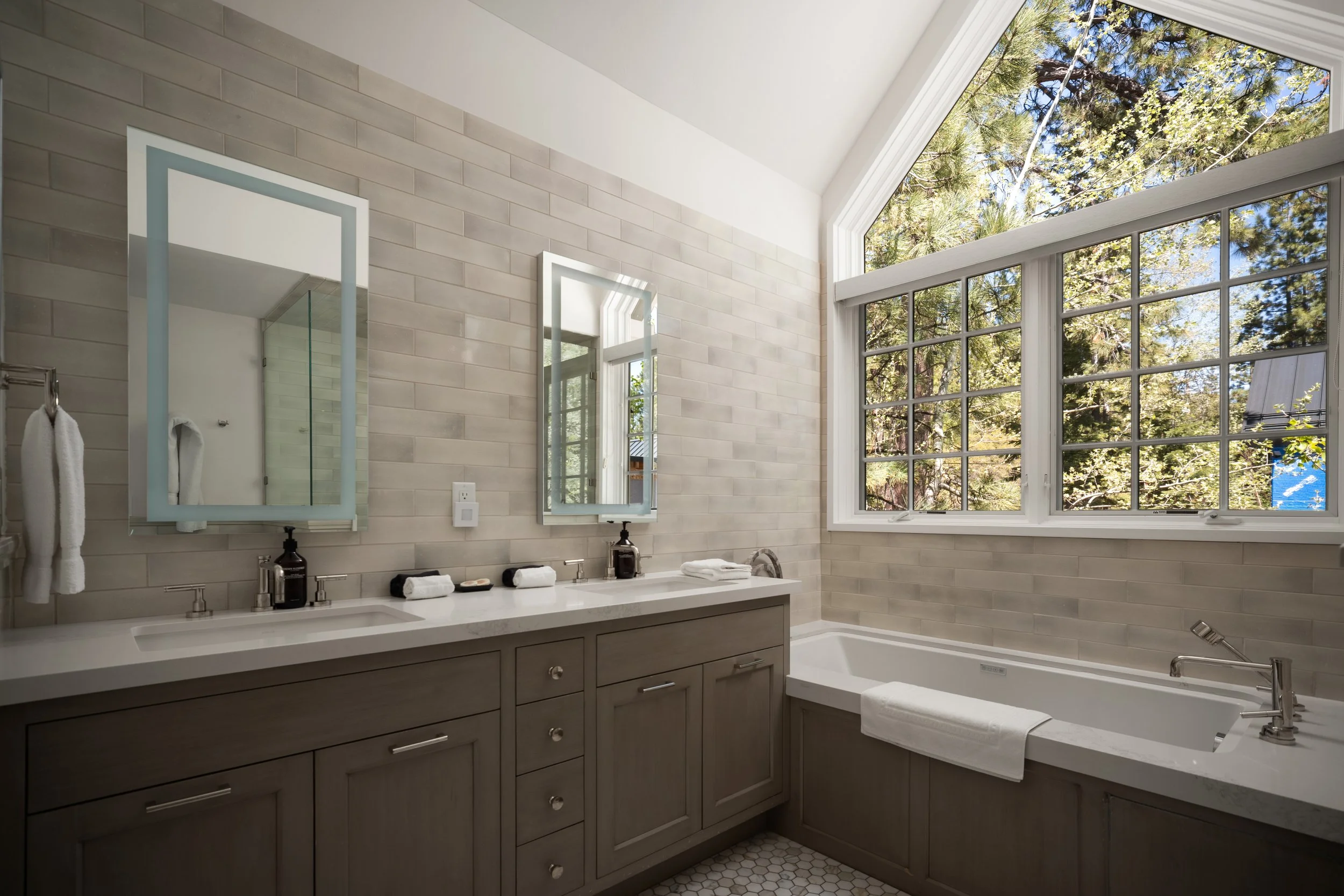 Modern bathroom with large window showing trees outside, double vanity with two mirrors and towels, and a bathtub.