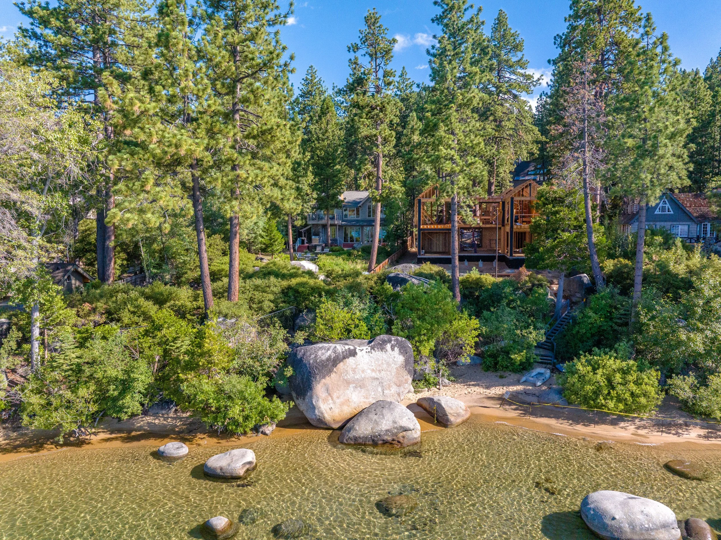 Scenic view of a lakeshore scene surrounded by tall pine trees and wooded houses under a blue sky with scattered clouds