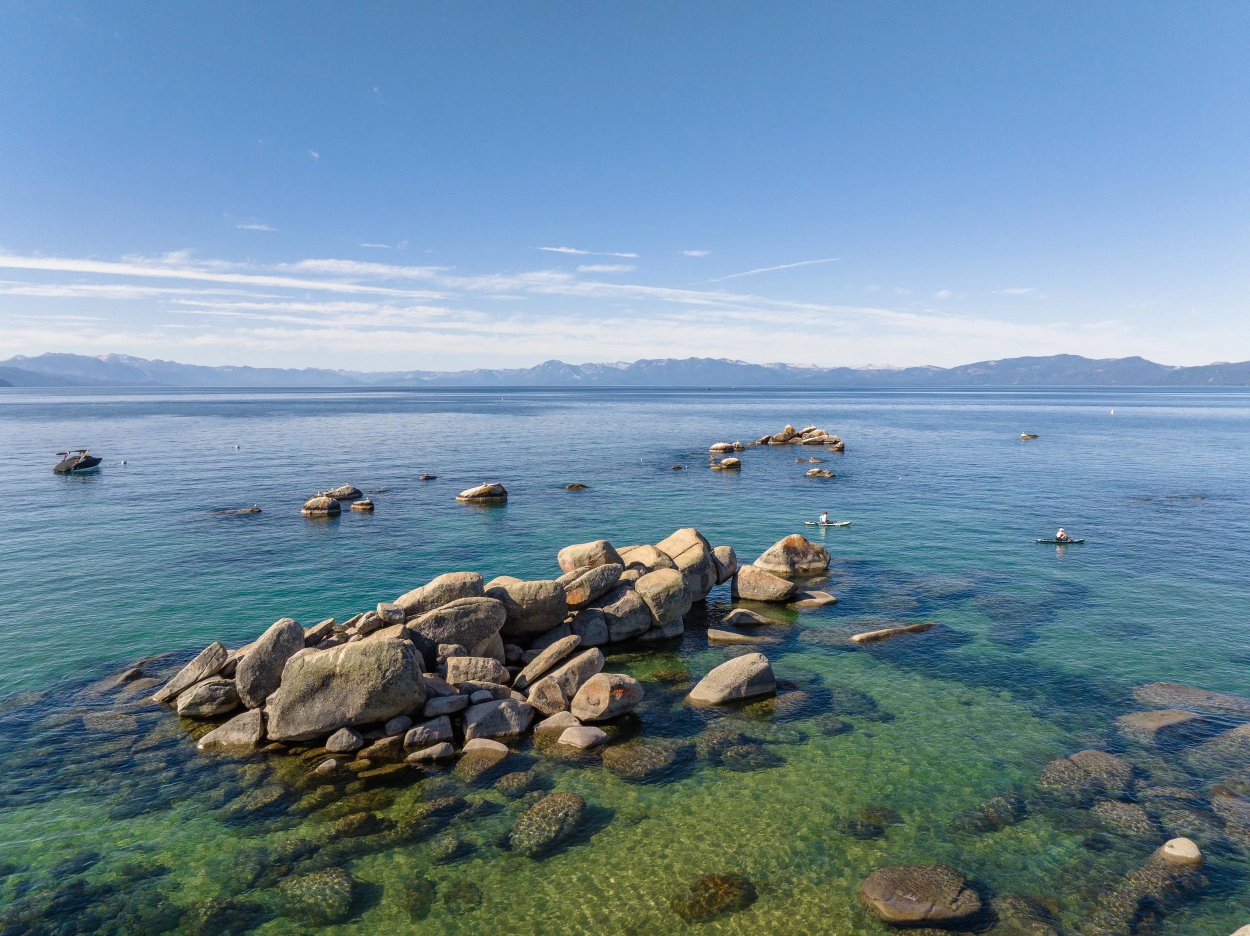 A scenic view of a calm body of water with rocks in the foreground, mountains in the distance, and a partly cloudy blue sky.