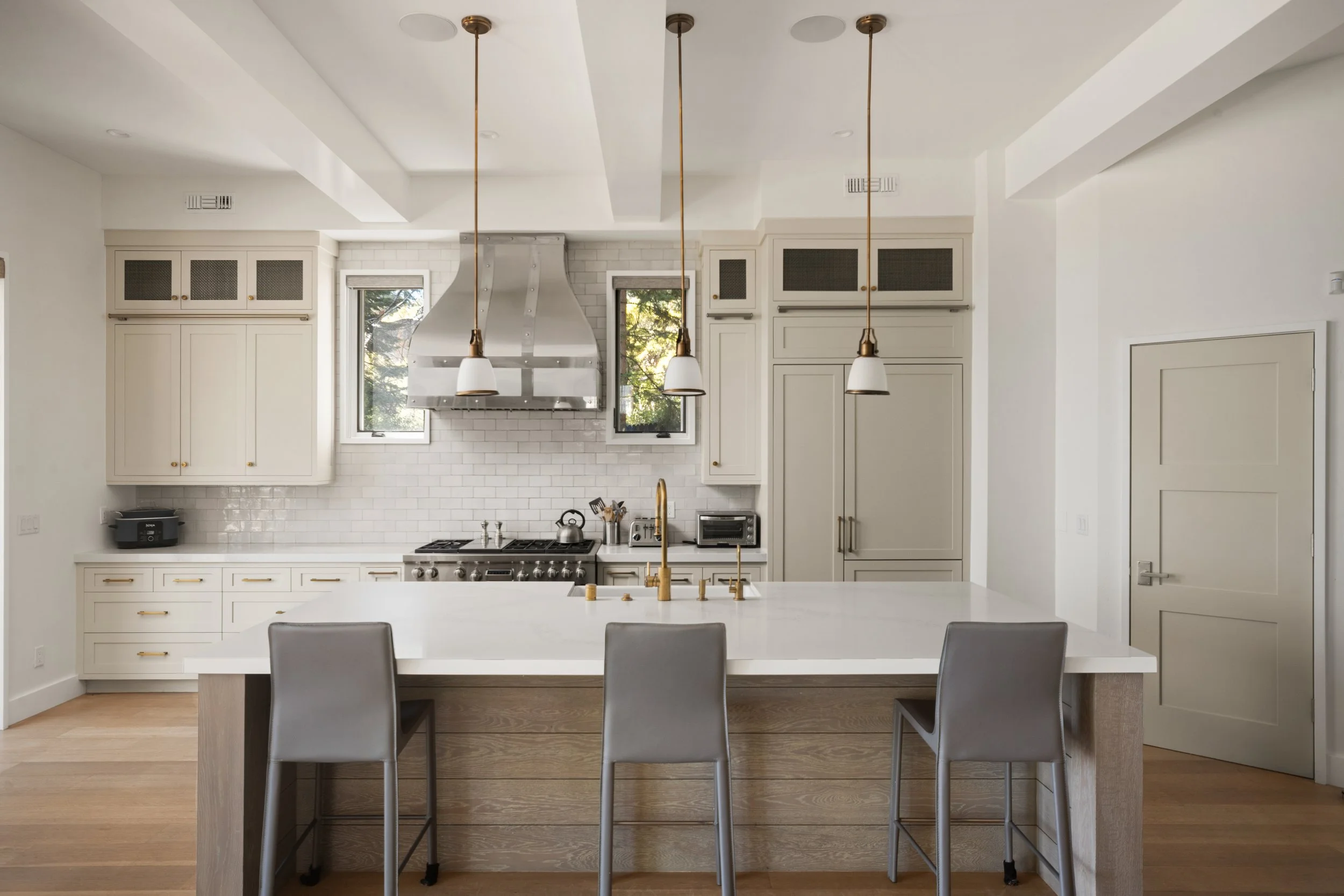 Modern kitchen with white cabinets, stainless steel range hood, stovetop, white subway tile backsplash, pendant lights, and a white island with gray chairs.
