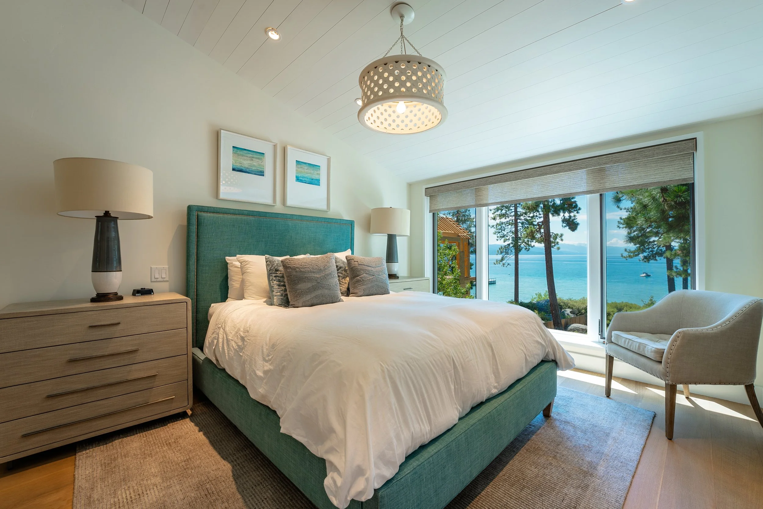 A bedroom with a large window showing trees and water outside. The room has a bed with white bedding, gray pillows, a teal upholstered headboard, wooden nightstands with lamps, and artwork above the bed. There is also a beige chair near the window.