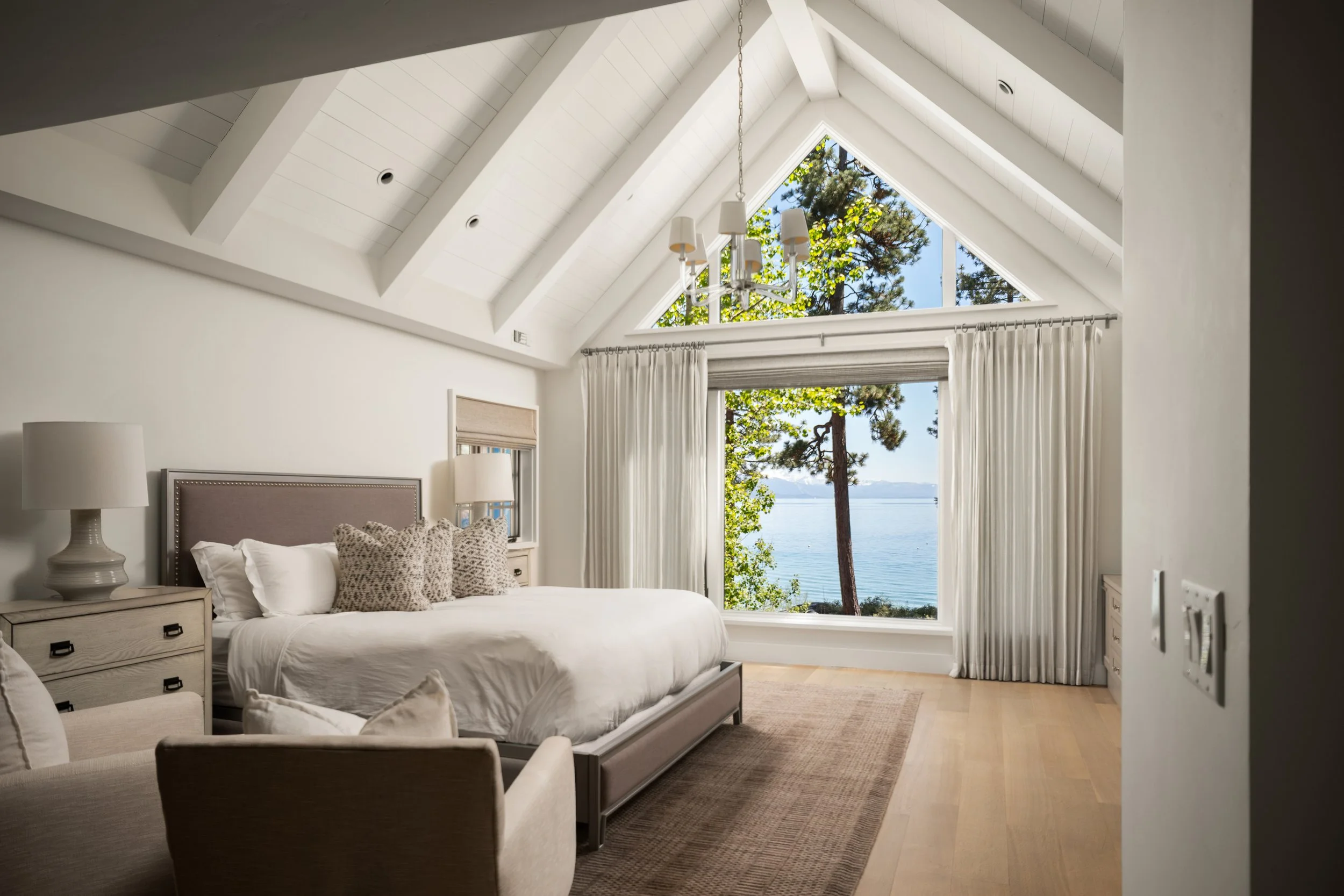 A bedroom with a large triangular window showcasing trees and water outside. The room features a white bed with patterned pillows, bedside tables with lamps, white curtains, and a chandelier hanging from the ceiling.