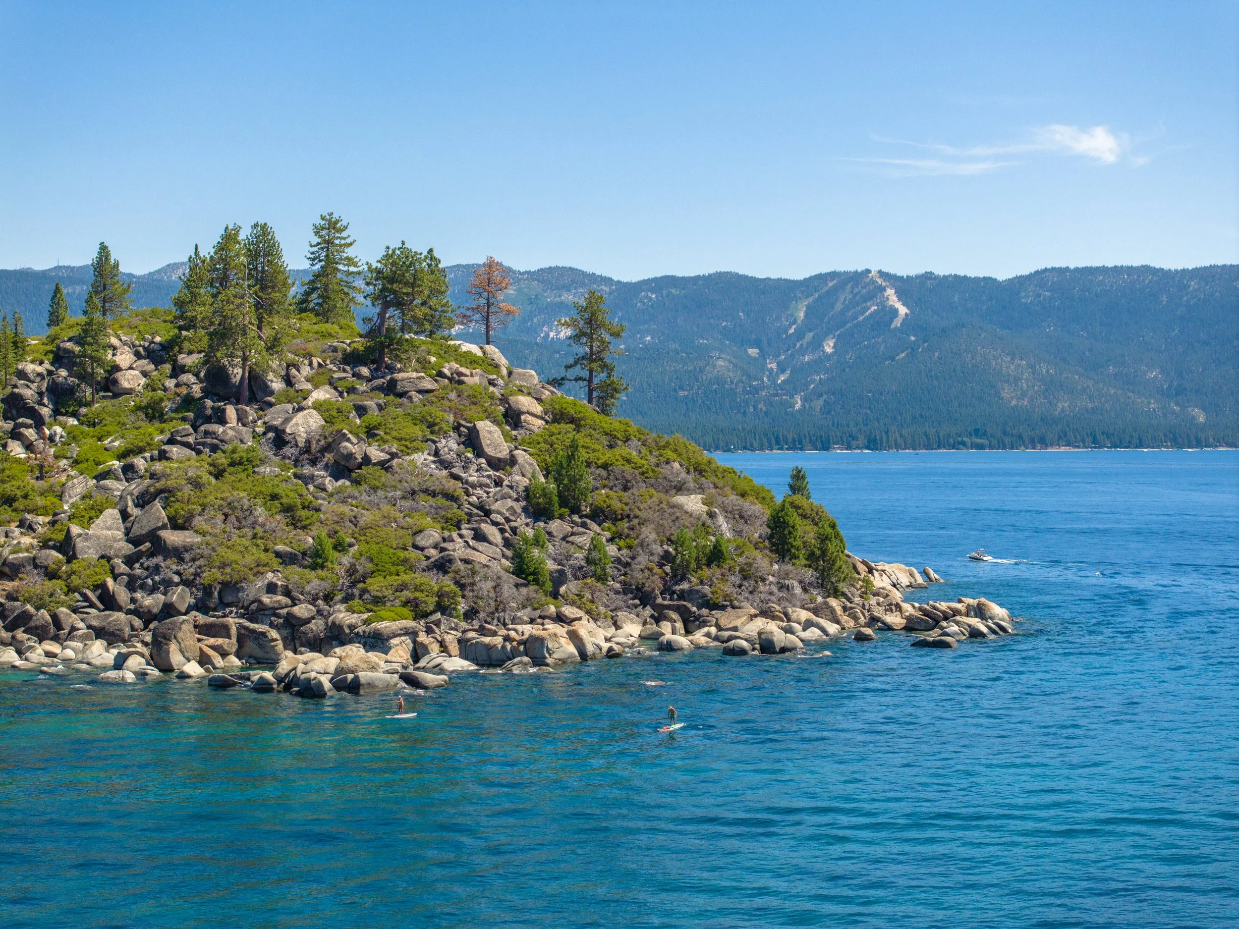 A scenic lakeside landscape with rocky shoreline, green trees, and mountain range under clear blue sky. People are paddleboarding on the water.