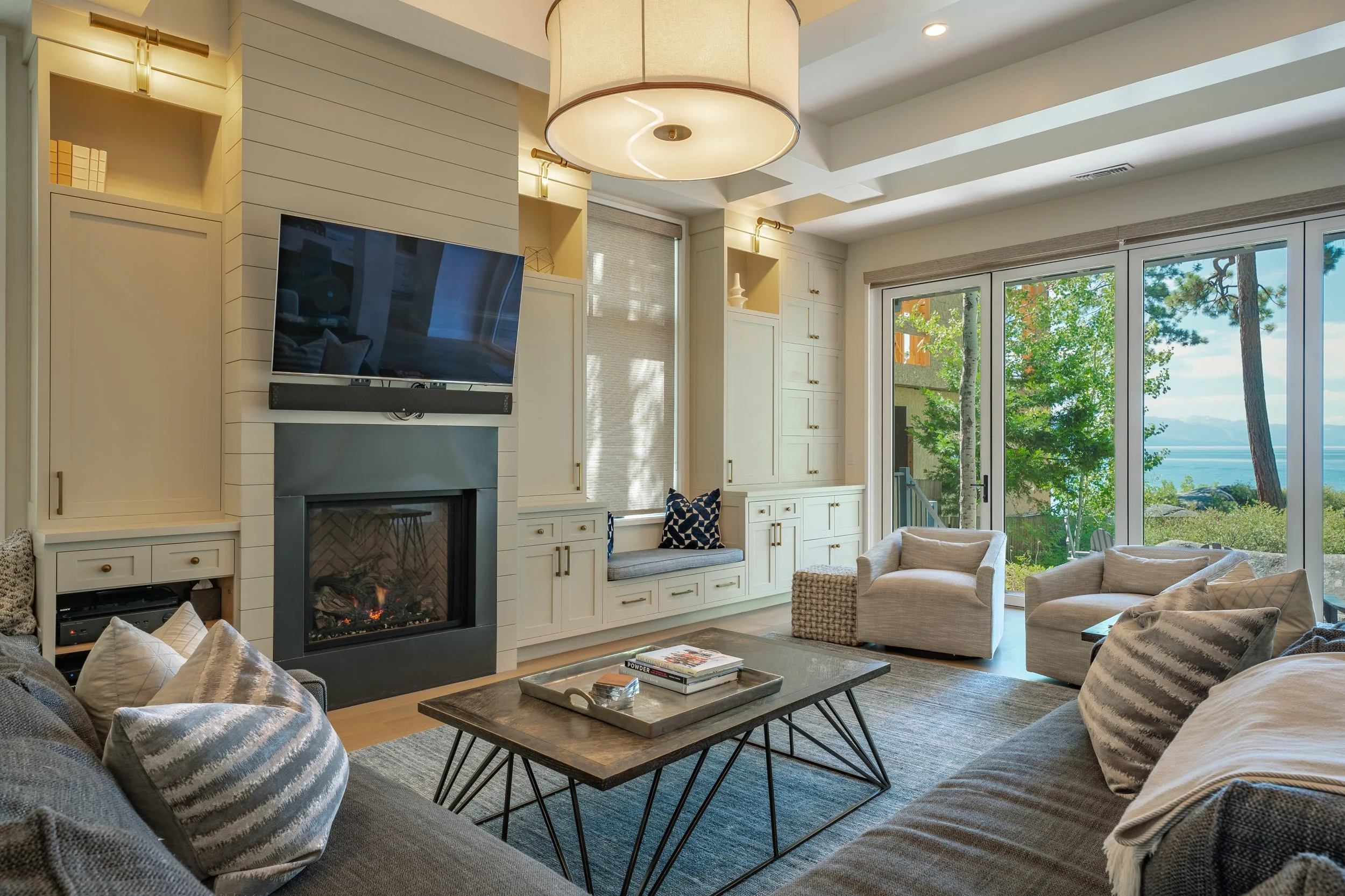 Living room with cream-colored cabinetry, a fireplace, a wall-mounted TV, large windows showing trees and water, and furniture including armchairs and a coffee table.