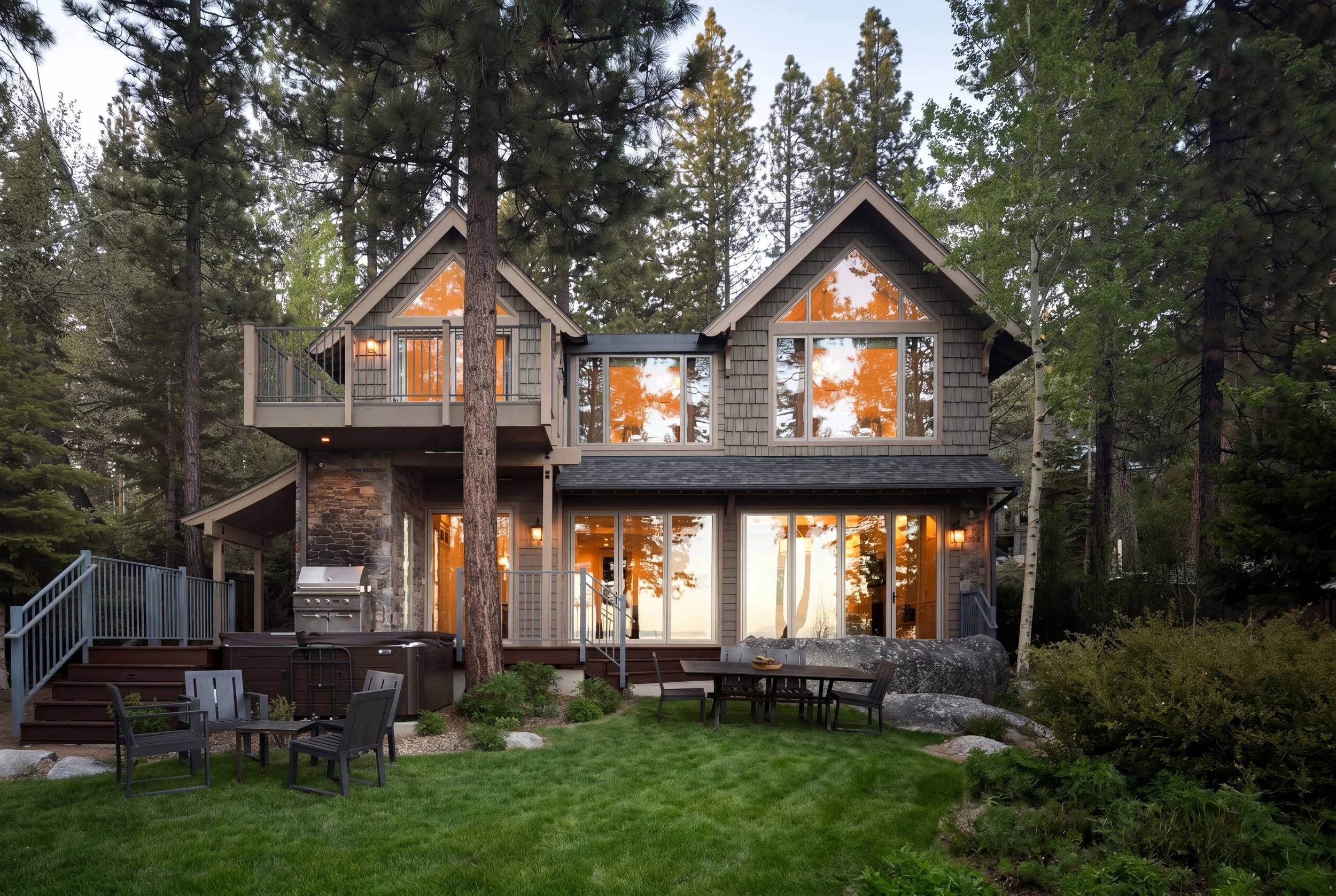 A modern house with large windows and a deck, surrounded by tall trees and greenery, during sunset.
