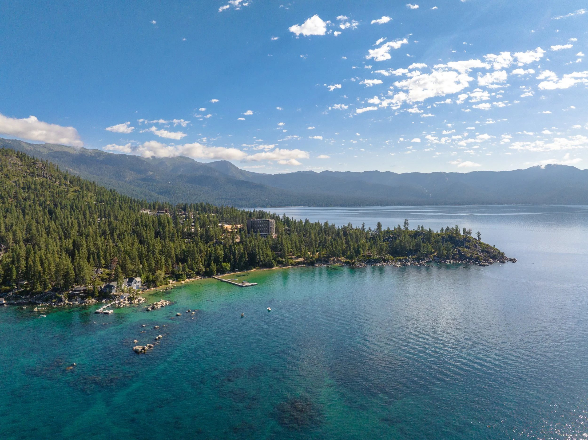 A scenic view of a calm lake with clear blue water, surrounded by a dense forest of green trees, and mountains in the background under a bright blue sky with scattered white clouds.