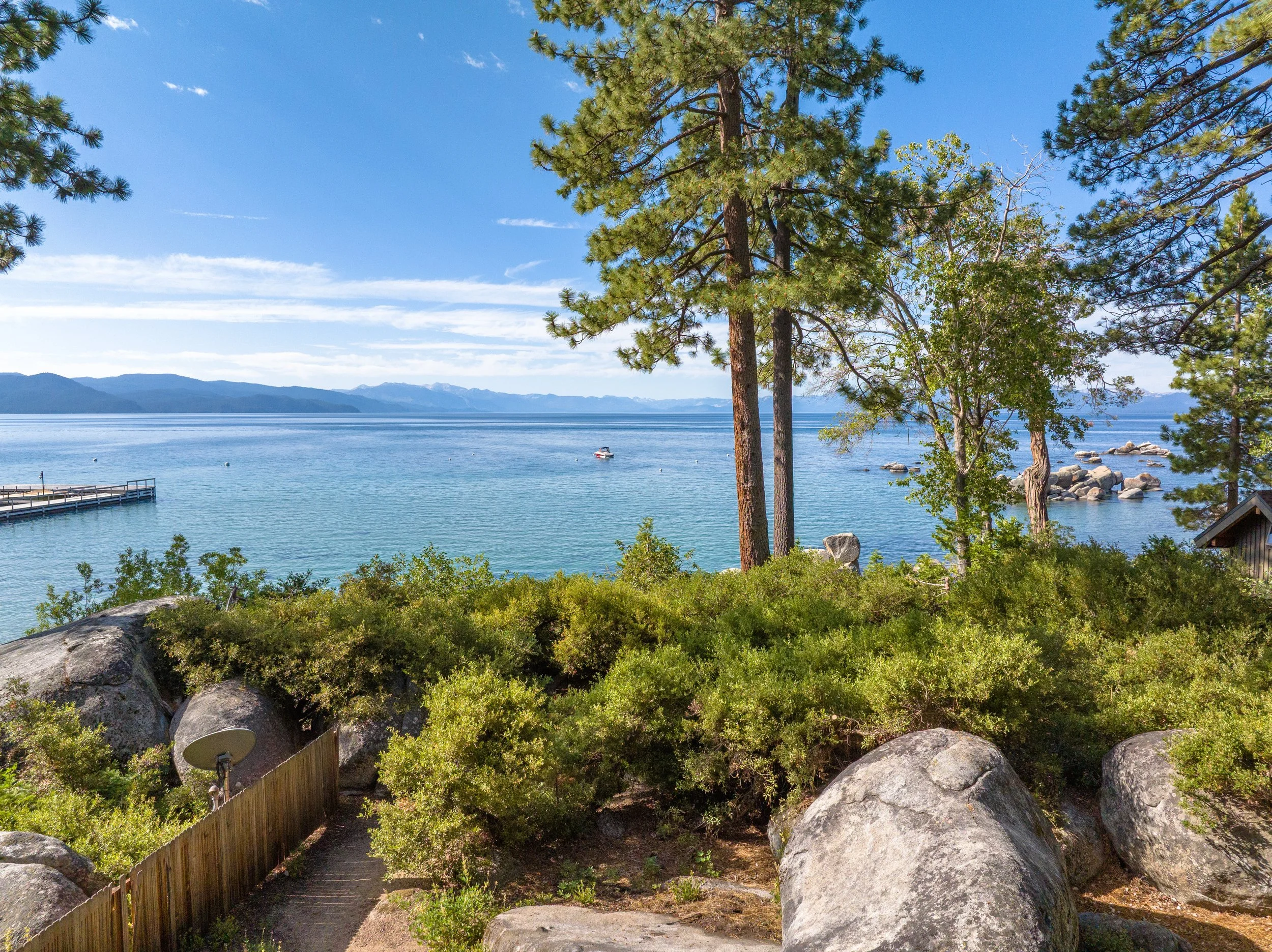 A view of a lake with mountains in the background, trees in the foreground, rocks along the shoreline, a small dock extending into the water, and a solitary boat on the lake under a clear blue sky with some clouds.
