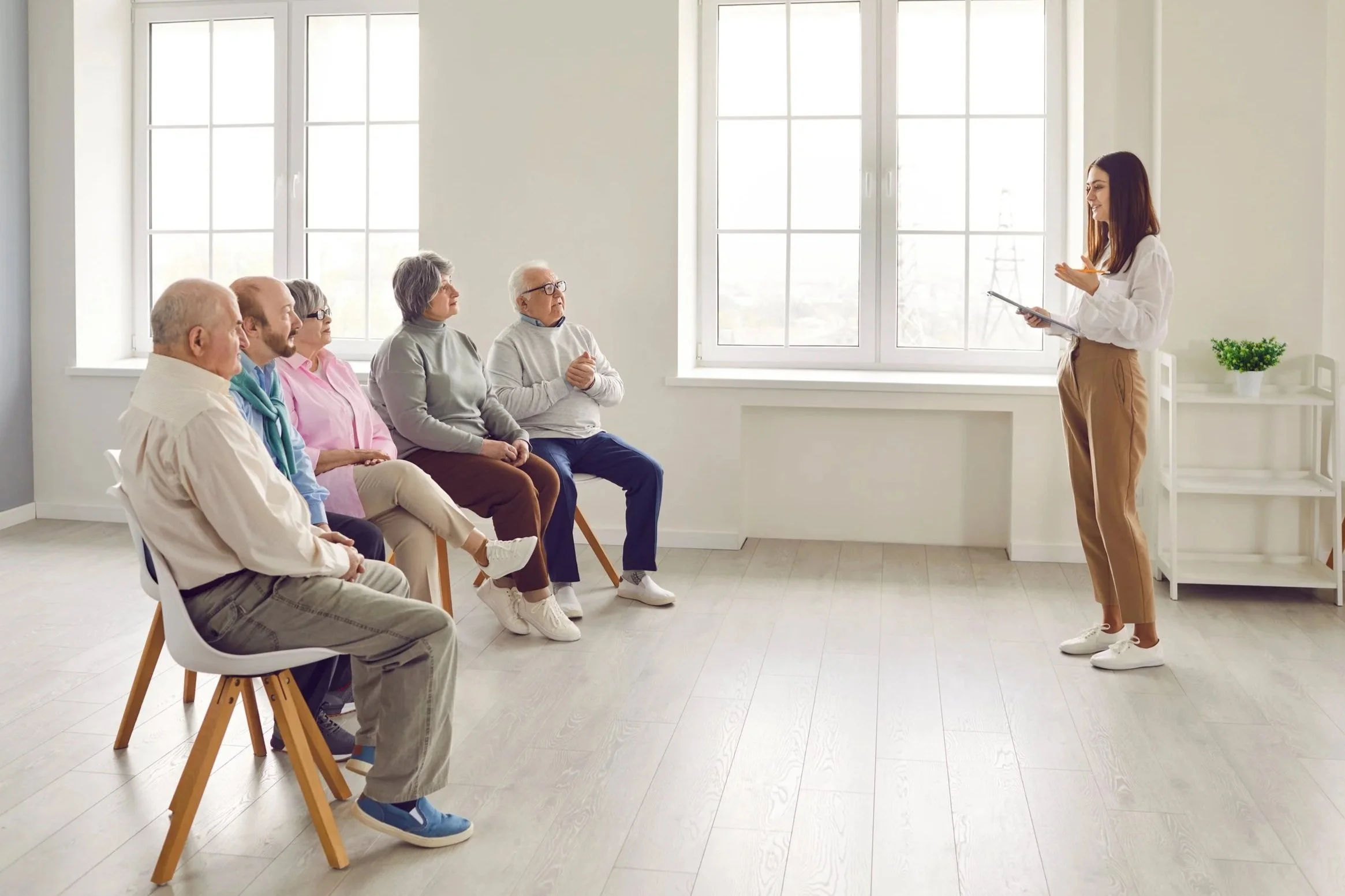 A young woman giving a presentation to an audience of five older adults in a bright, minimalist room with large windows. The woman is holding a tablet and appears to be speaking to the seated group.