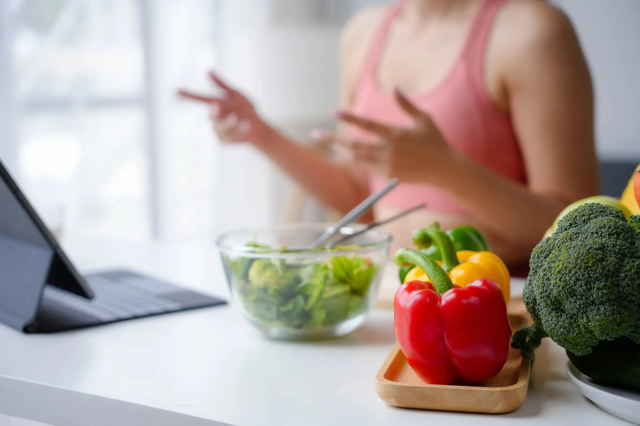 Fresh vegetables, including red and yellow bell peppers, broccoli, and a bowl of salad, on a kitchen counter. A woman wearing a pink sleeveless top is in the background, preparing food.