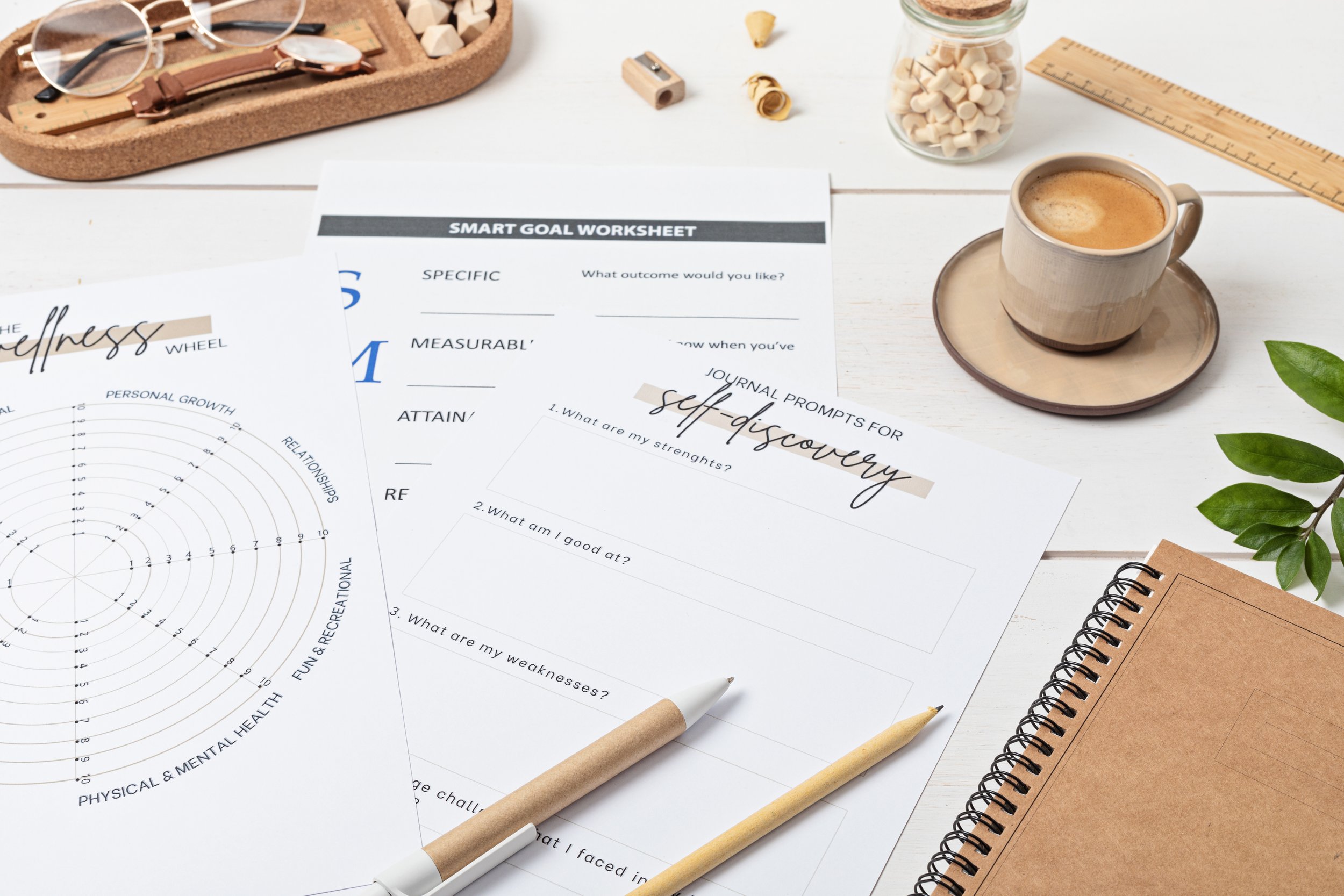 A workspace featuring a coffee mug on a saucer, various worksheets including a SMART goal worksheet and a self-discovery worksheet, a spiral notebook, pens, a ruler, and a jar of white push pins, with some green leaves visible on the right side.