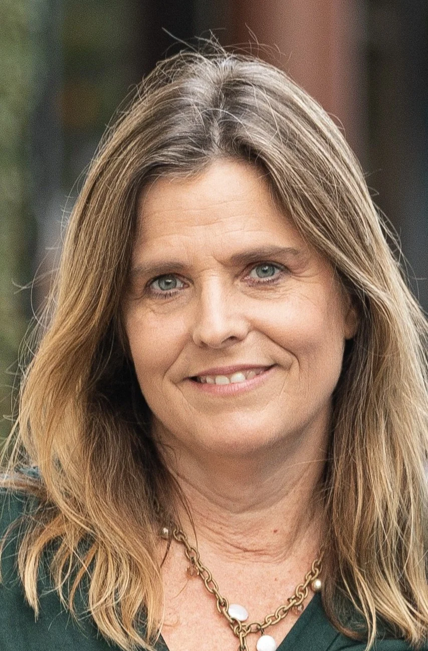 Close-up portrait of a woman with shoulder-length blonde hair, blue eyes, and a warm smile, wearing a green top and a gold necklace with pearls.