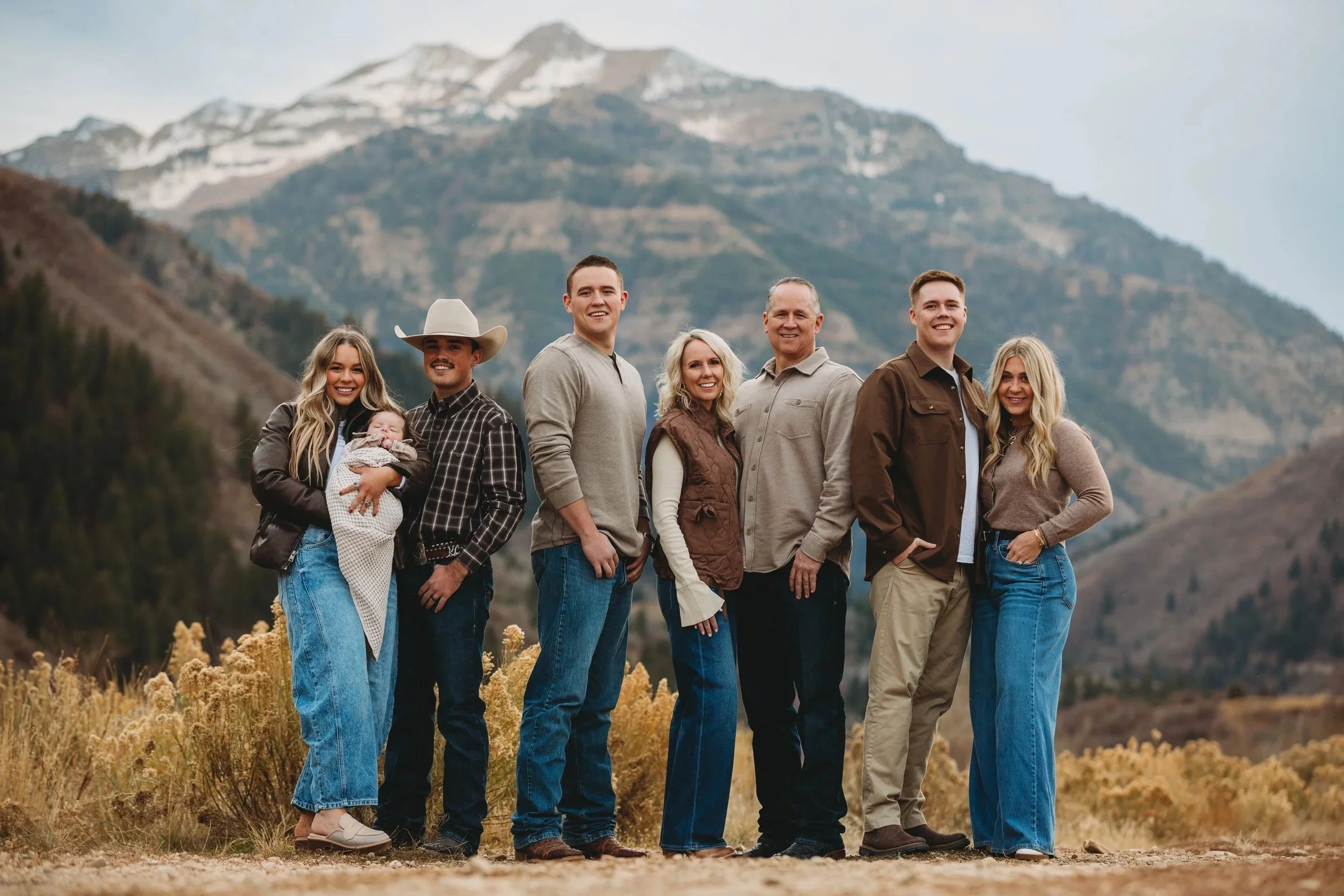 A multi-generational family of eight people standing outdoors in front of a mountainous landscape with snow-capped peaks, wearing casual clothing and smiling at the camera.