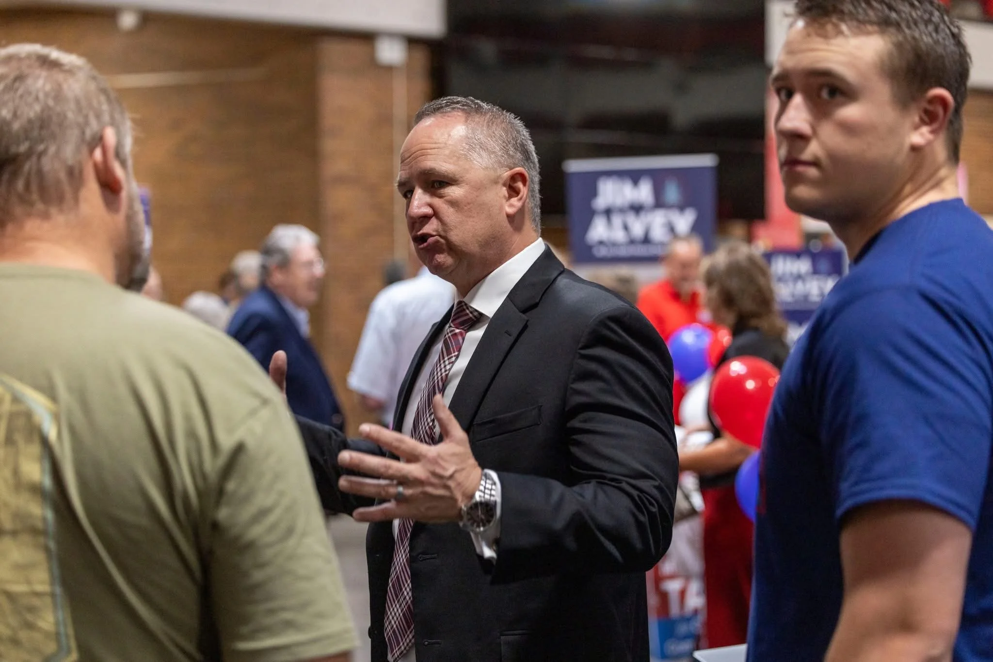 Three men engaged in conversation at a political event, with signs in the background supporting Juma Alvey.