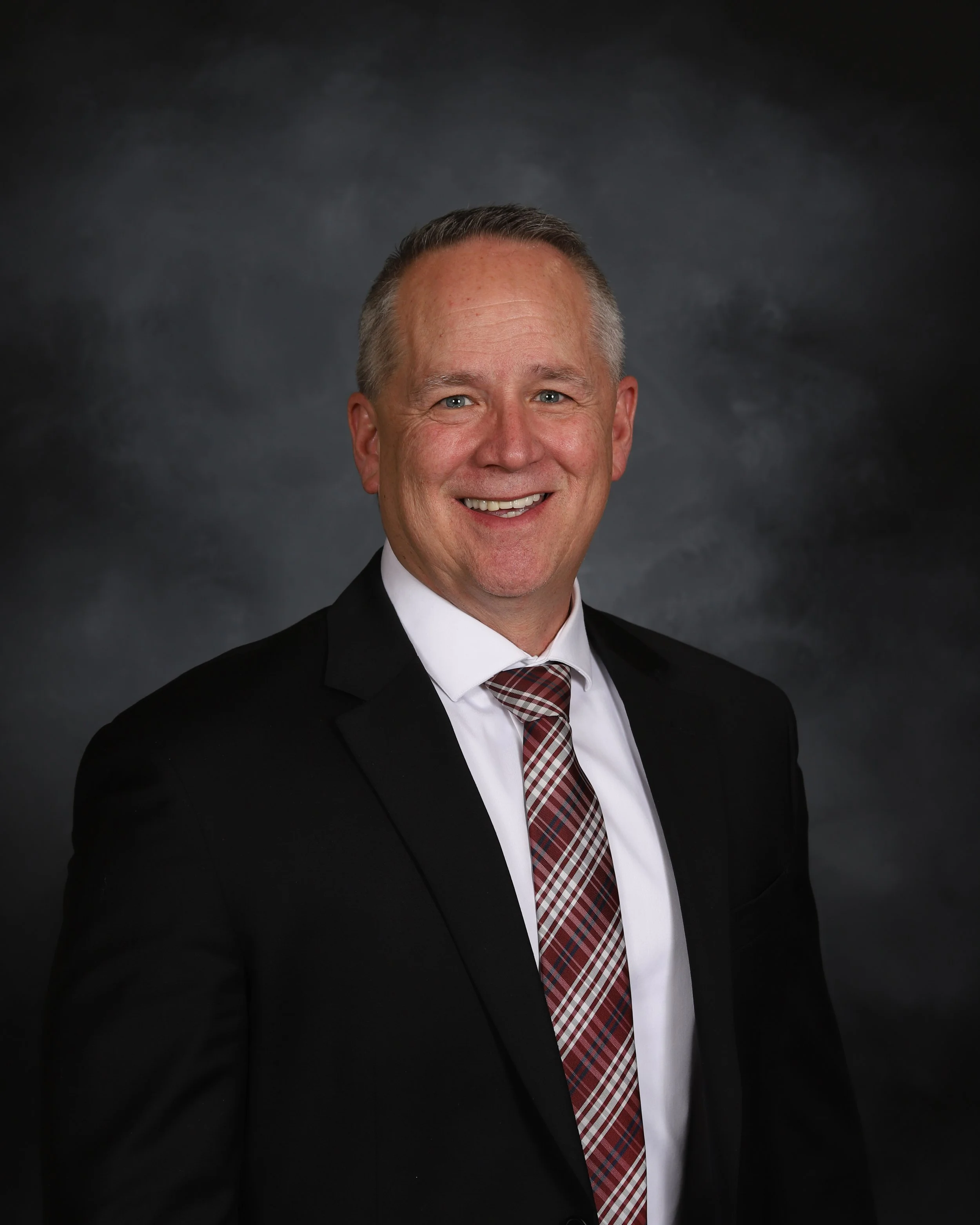 Portrait of a smiling middle-aged man with short gray hair, dressed in a black suit, white shirt, and a red plaid tie against a dark background.