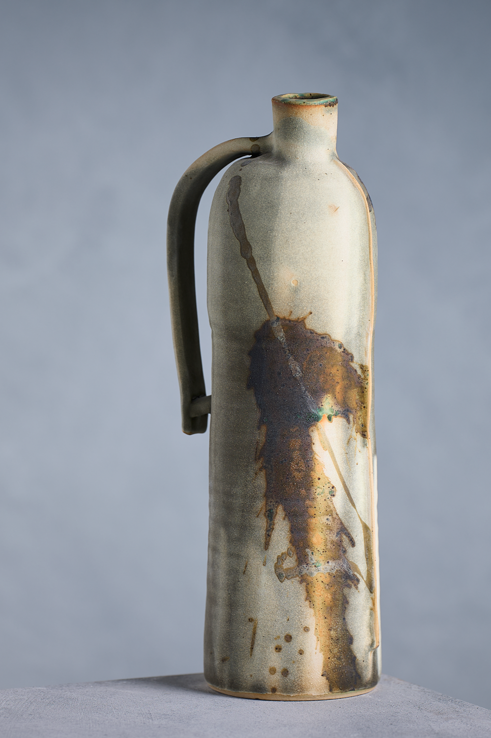 A ceramic bottle with a long neck, rounded handle, and glaze that resembles a burnt or rusted leaf pattern.