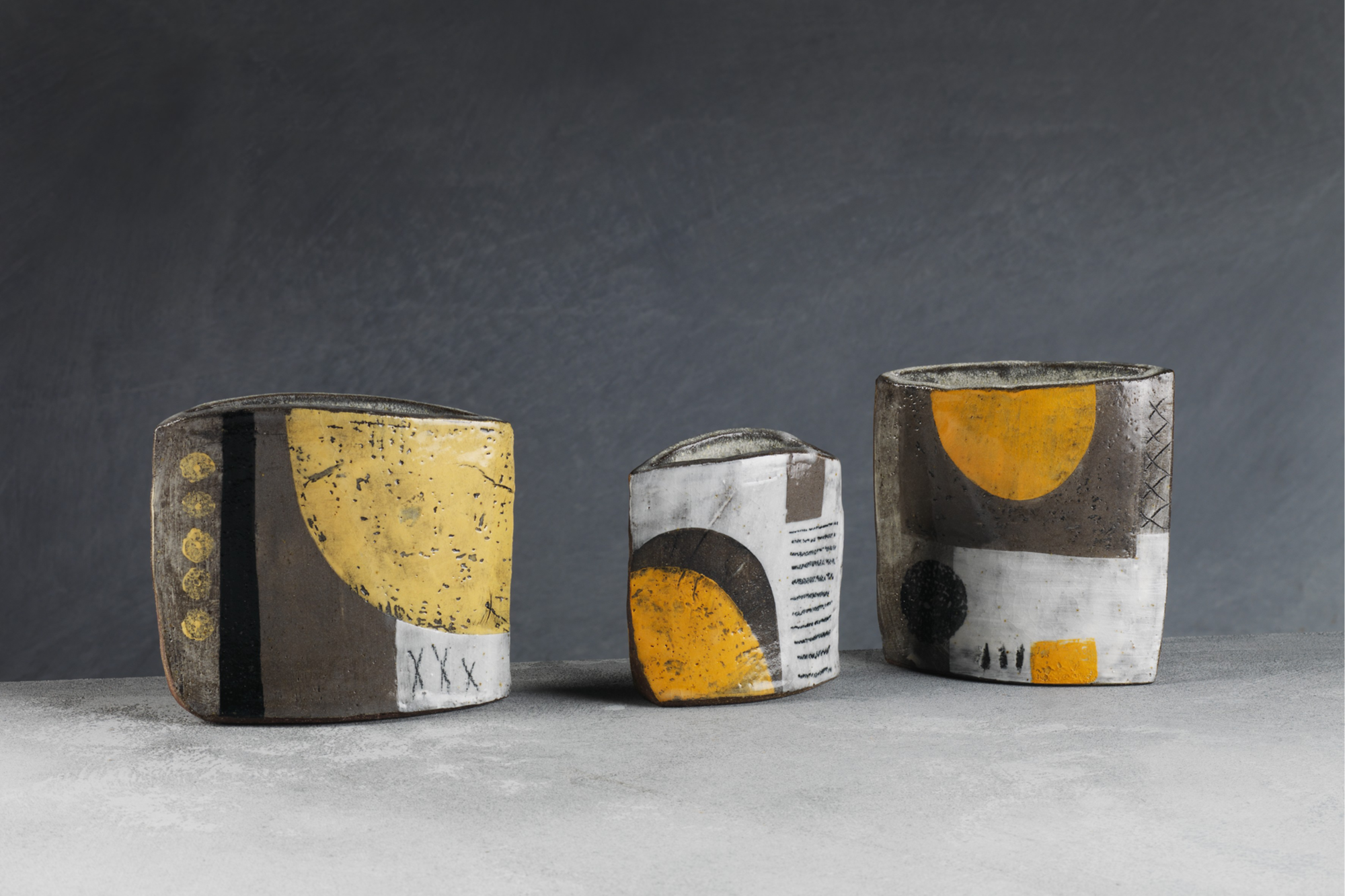 Three small ceramic sculptures with geometric patterns in yellow, black, white, and gray colors positioned on a gray surface against a dark gray background.