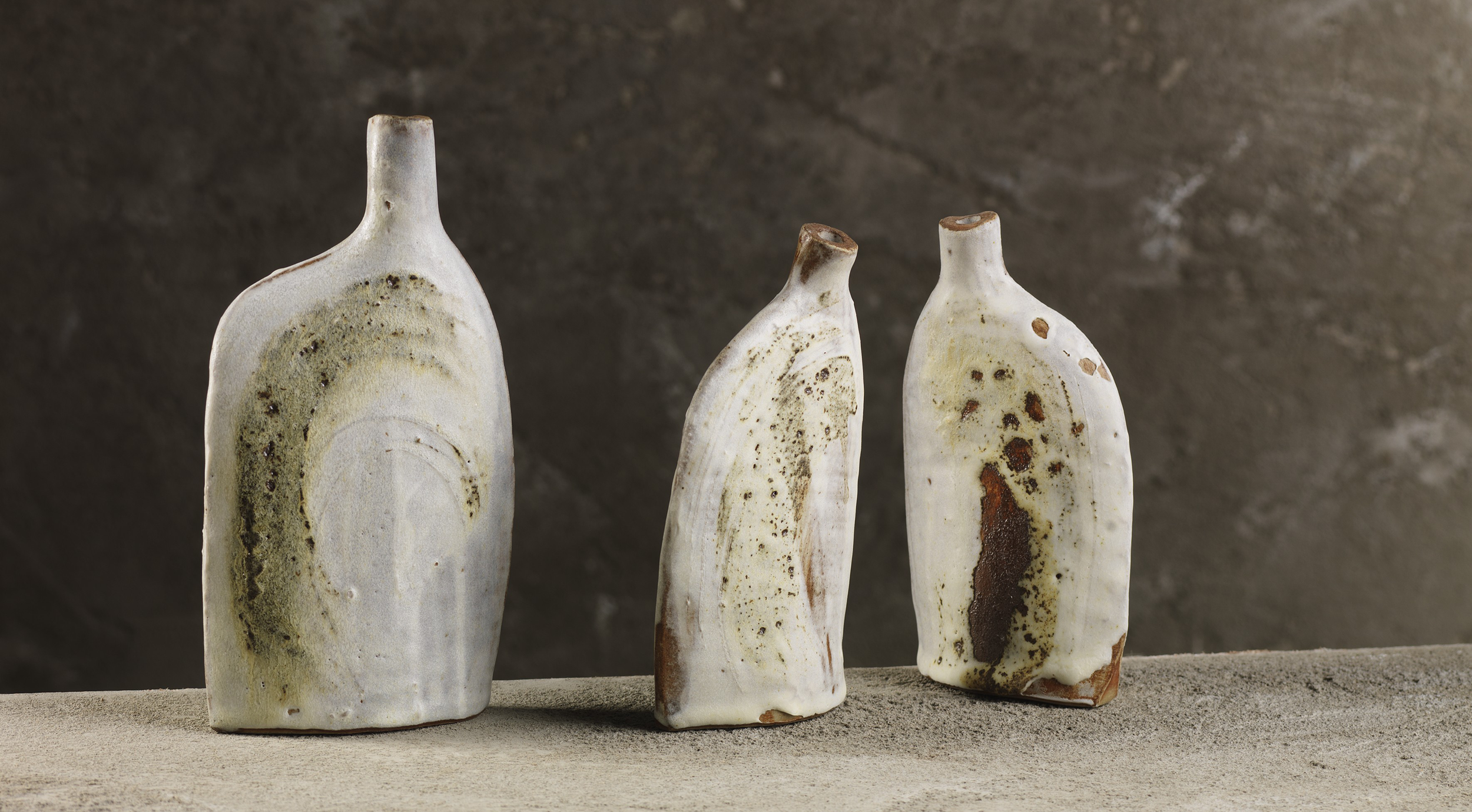 Three ceramic vases with earthy glaze finish, standing on a sandy surface against a concrete wall background.