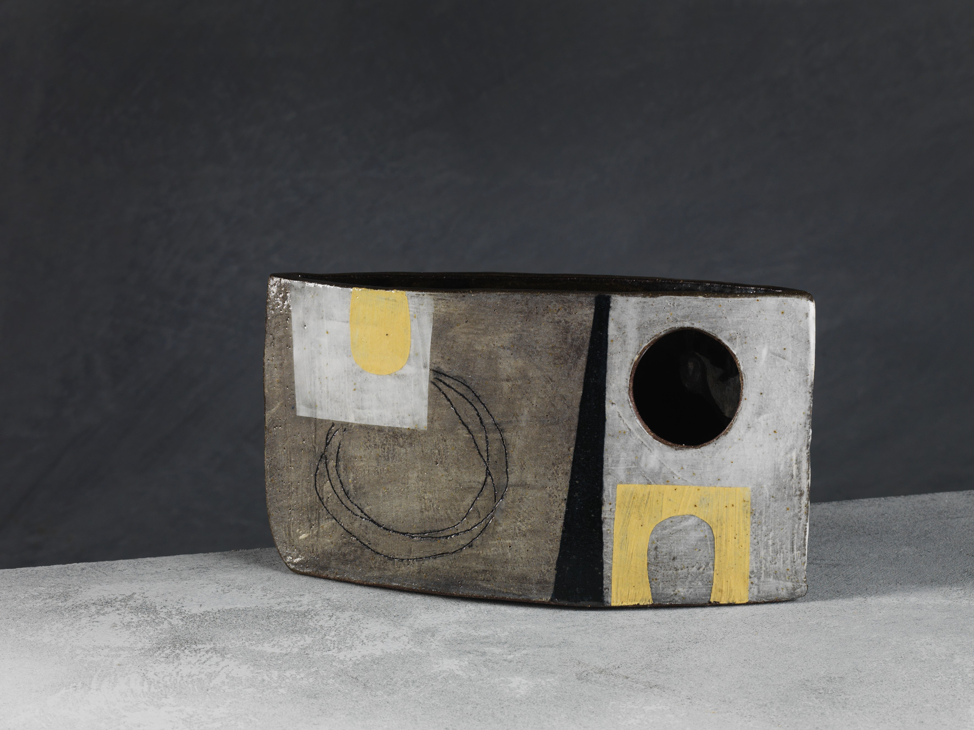 A rectangular ceramic sculpture with abstract geometric designs, including a black circle, black lines, and a yellow square patch, set against a dark background.