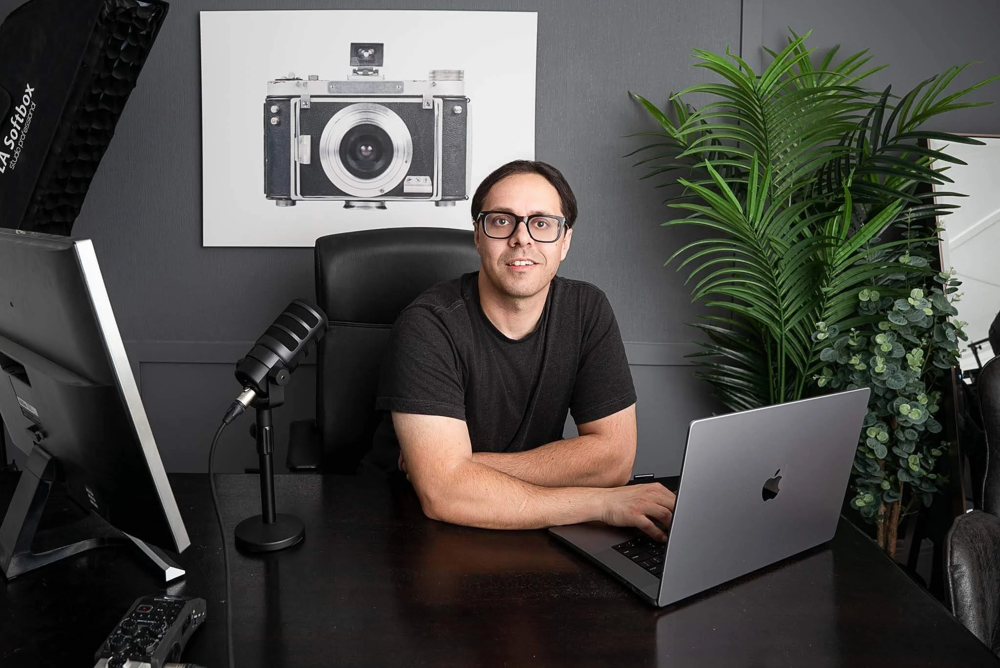 A man with glasses sits at a black desk with a silver MacBook, a large monitor, a microphone, and audio equipment. Behind him, there is a black and white framed photo of a vintage camera on a gray wall and a tall green plant to his right.