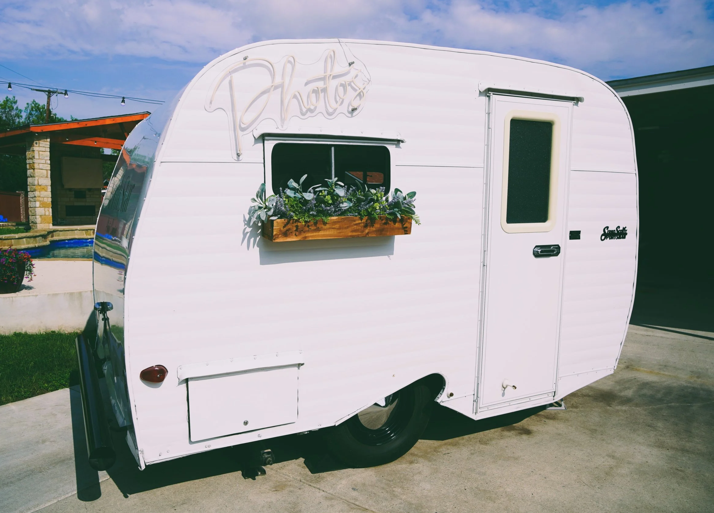 White travel trailer photo booth with a sign reading 'Photos' and a small window, parked on a concrete driveway with grass and trees in the background.