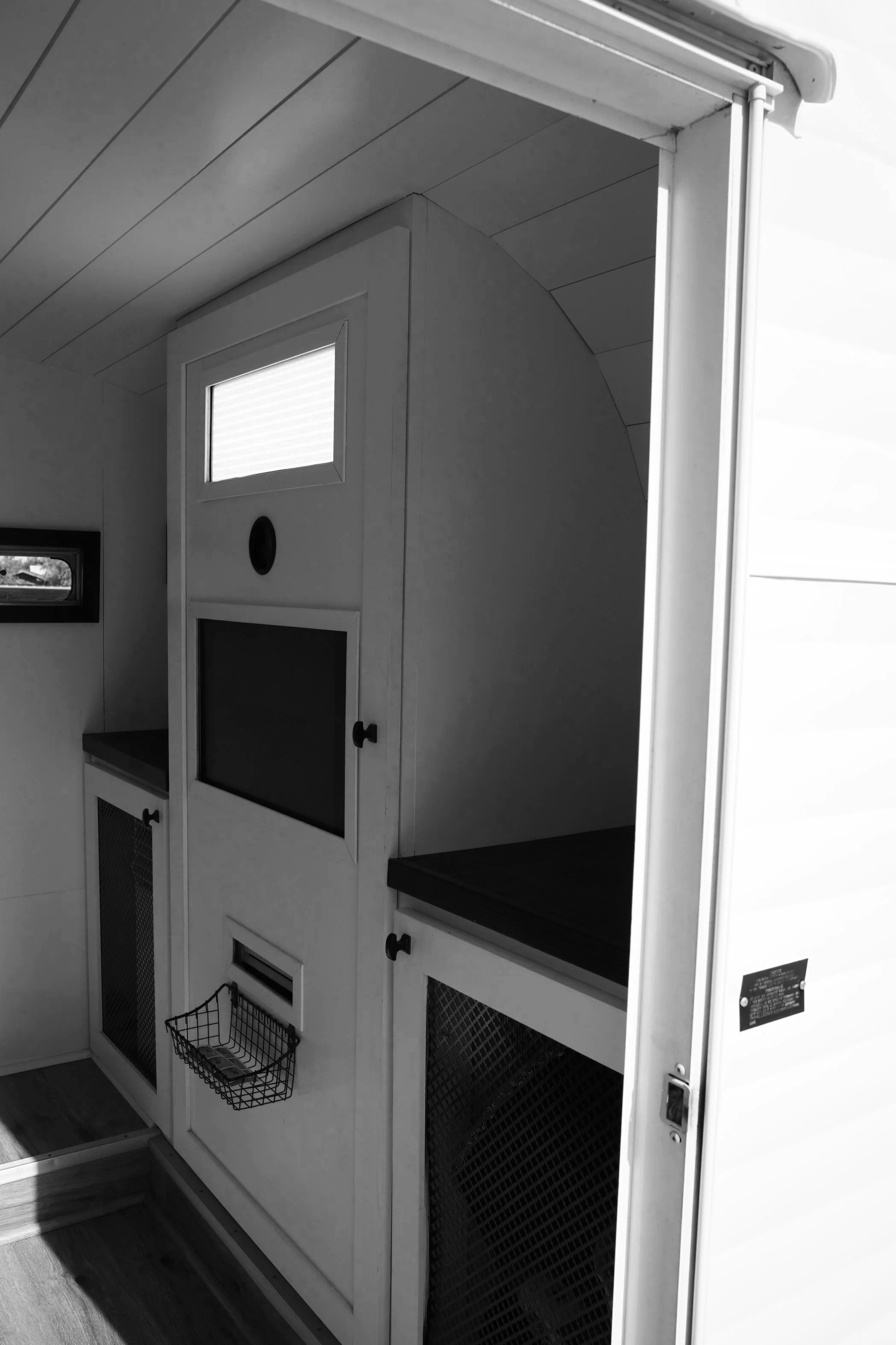 Black and white photo of the inside of a photo booth camper with multiple sections, including a small window on the upper part, a black chalkboard space, and wire baskets below, against a wall with wood flooring.