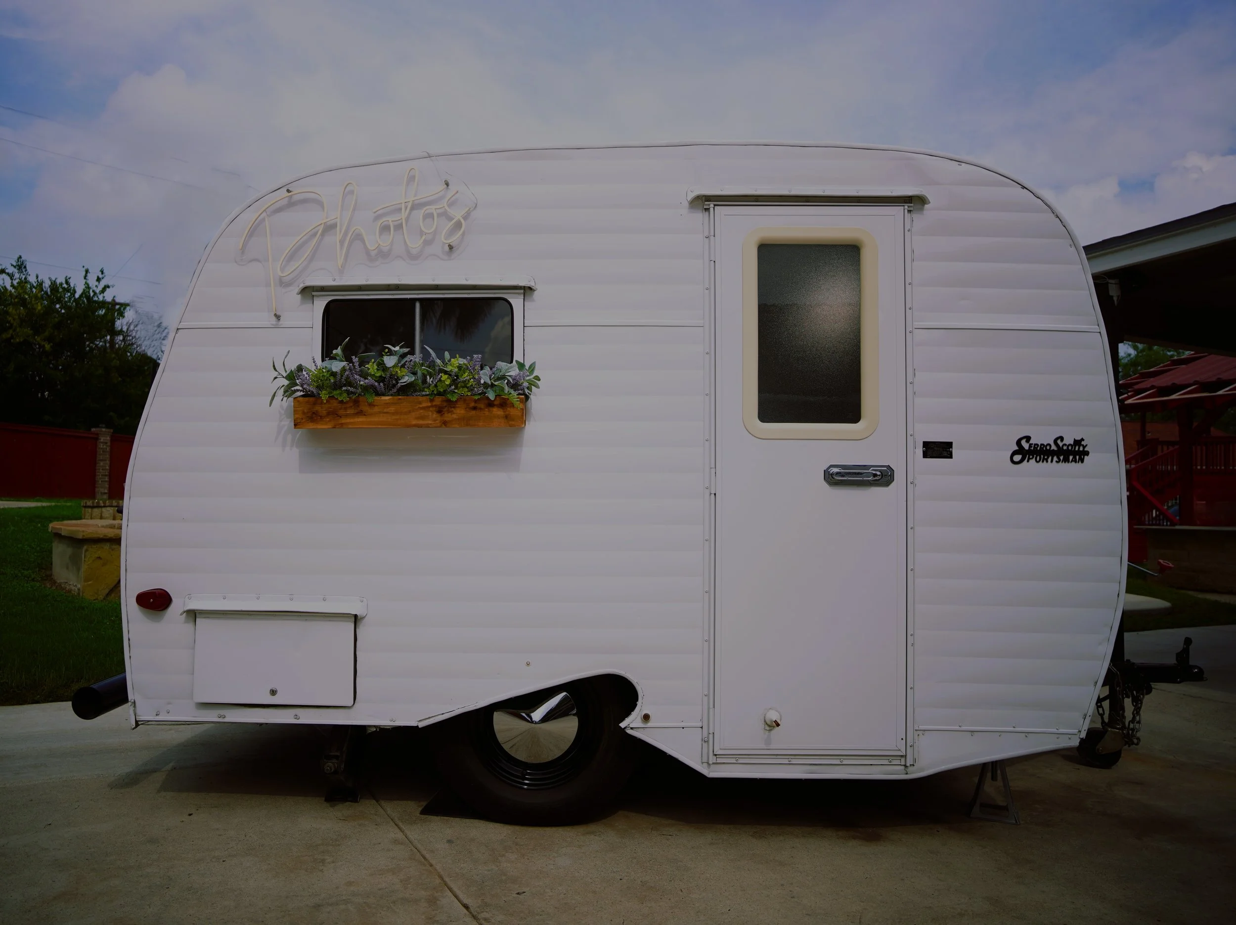 A white vintage photo boothcamper trailer parked on concrete ground under a partly cloudy sky. It has a small window with a flower box containing purple and green plants, a door with frosted glass, and a neon sign spelling 'Photos' on the upper side.