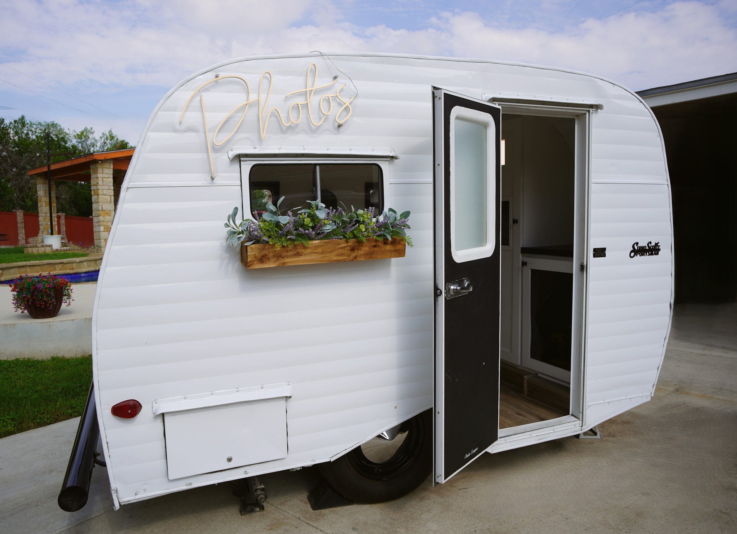 White photo booth camper trailer with a neon sign that says 'Photos' on the front, a small window with a flower box, and an open door revealing the interior, parked on a concrete driveway with a house and trees in the background.