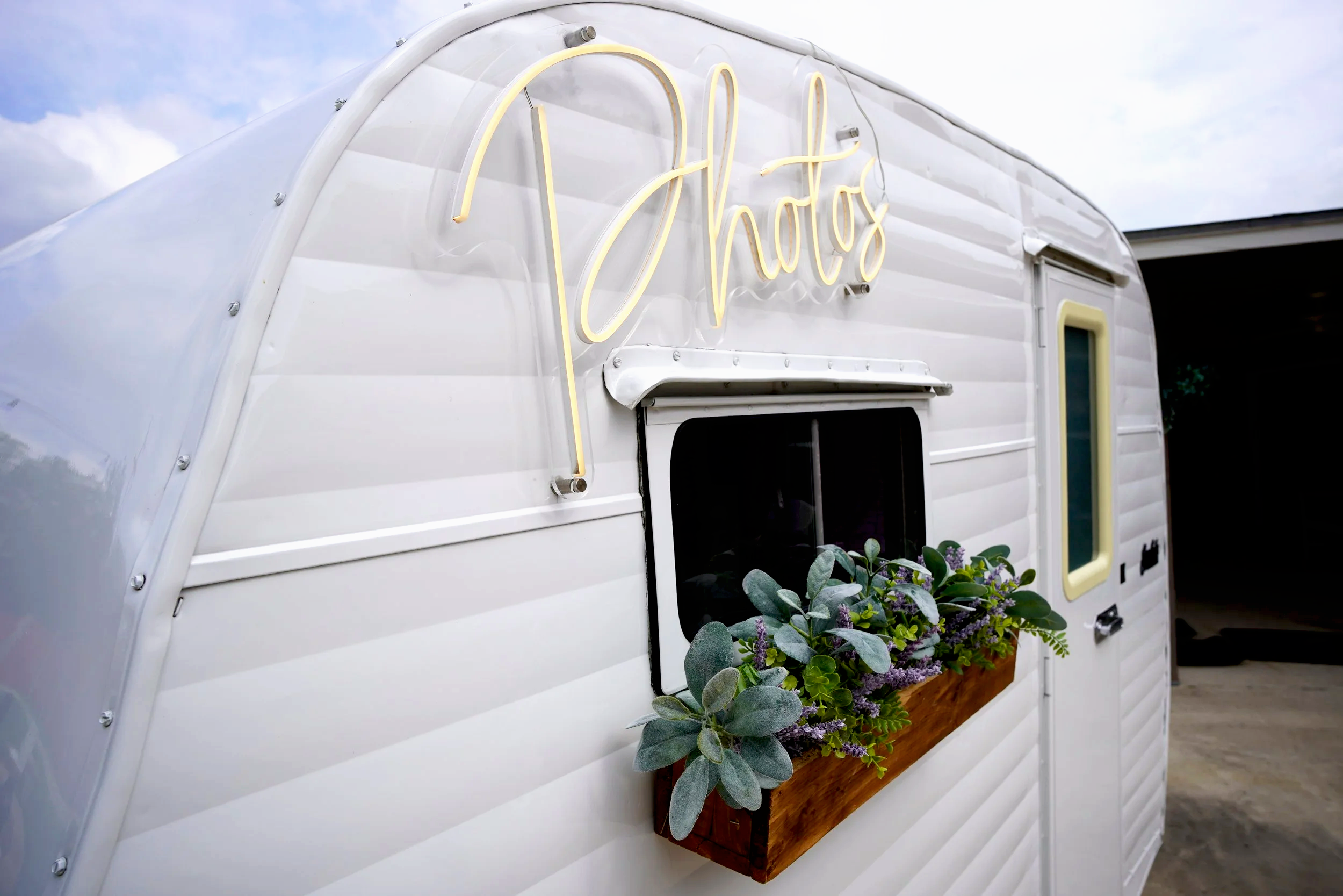 A white vintage photo booth camper with a neon yellow sign that says ''Photos,'' a window, and a flower box with green plants and purple flowers on the side.