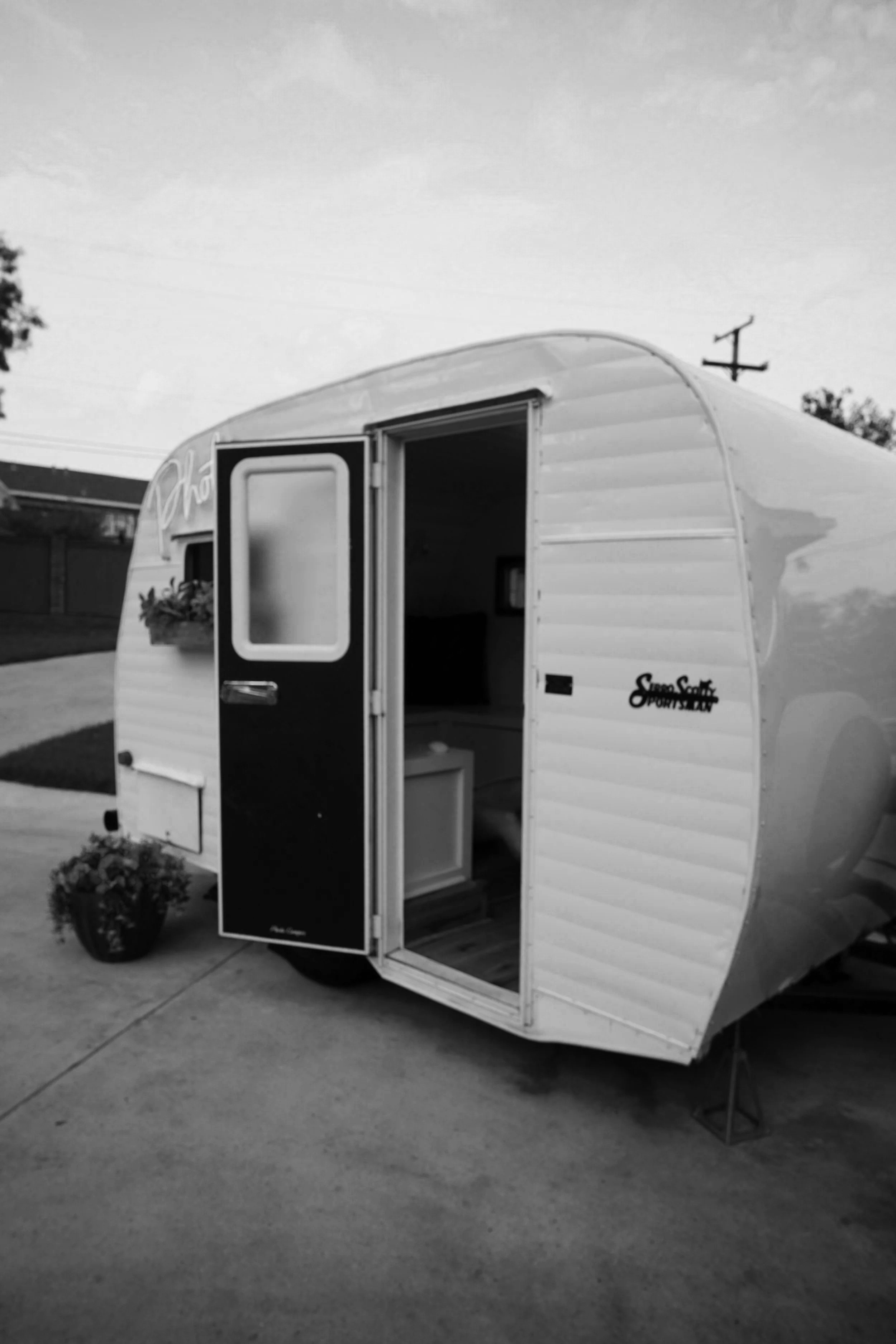 A small photo booth camper with an open door, parked on a concrete driveway with a flower pot nearby, and power lines and trees in the background.