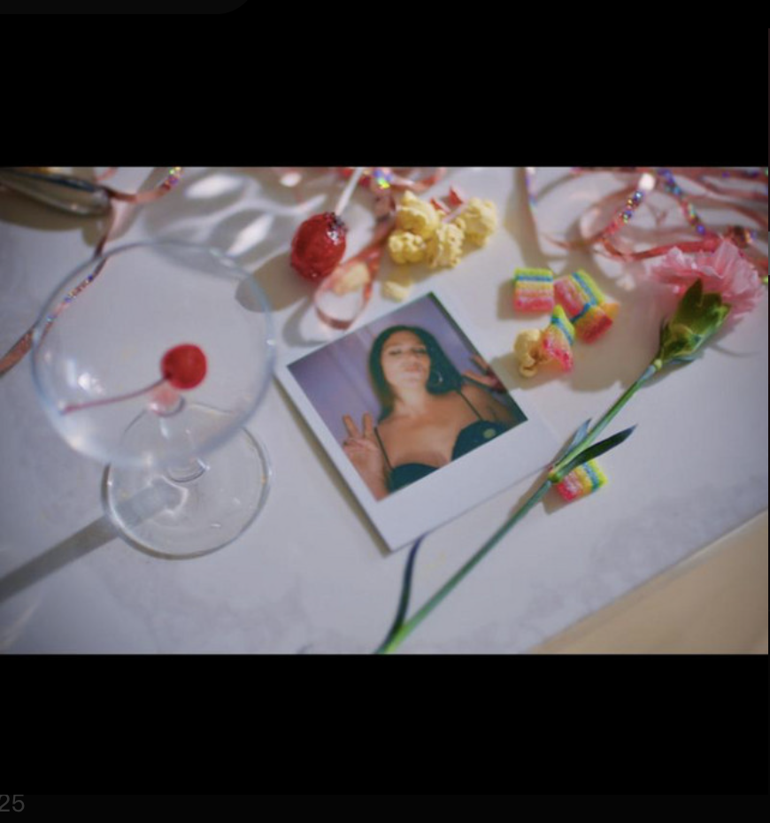 A table decorated with colorful candies, a pink flower, a photo of a woman making a peace sign, and pink streamers, suggesting a celebration or party.